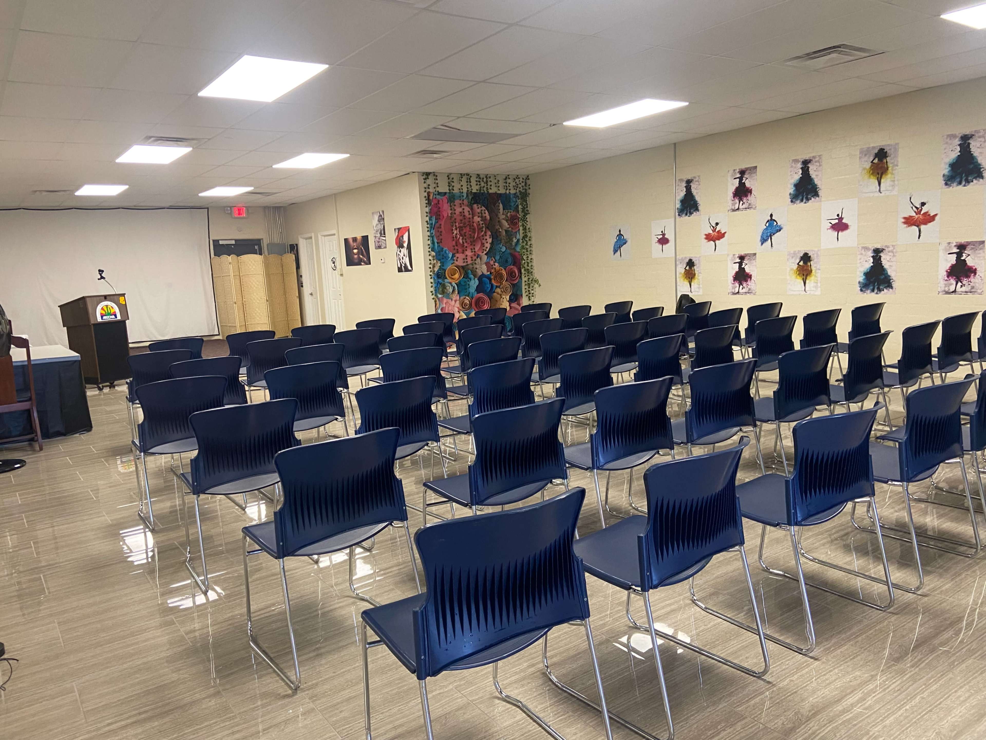 A room arranged with rows of blue chairs facing a presentation area, with colorful artwork displayed on the walls.