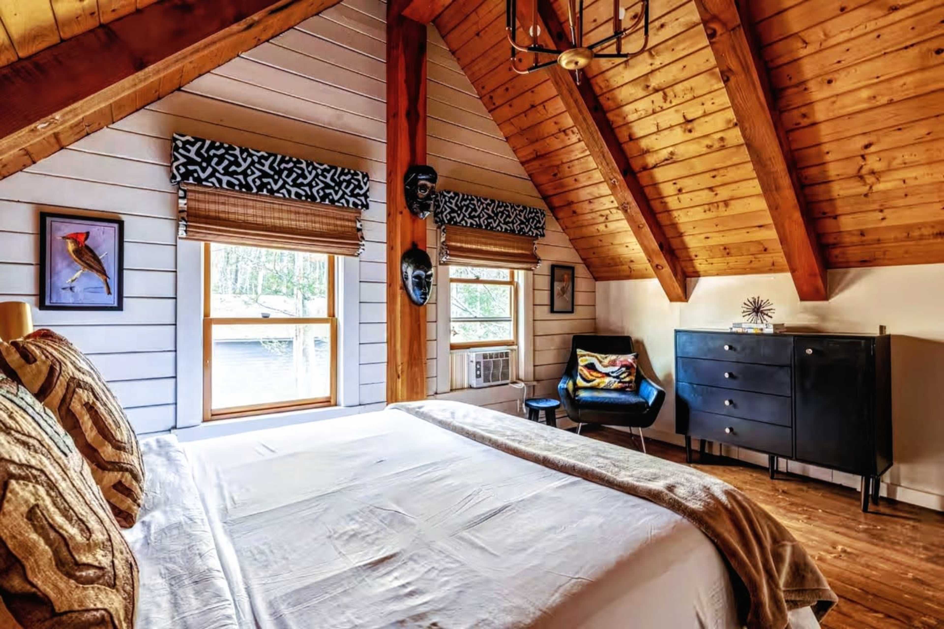 The image depicts a cozy bedroom with a pitched wooden ceiling, featuring a large bed, two windows, a black dresser, and a chair in the corner.