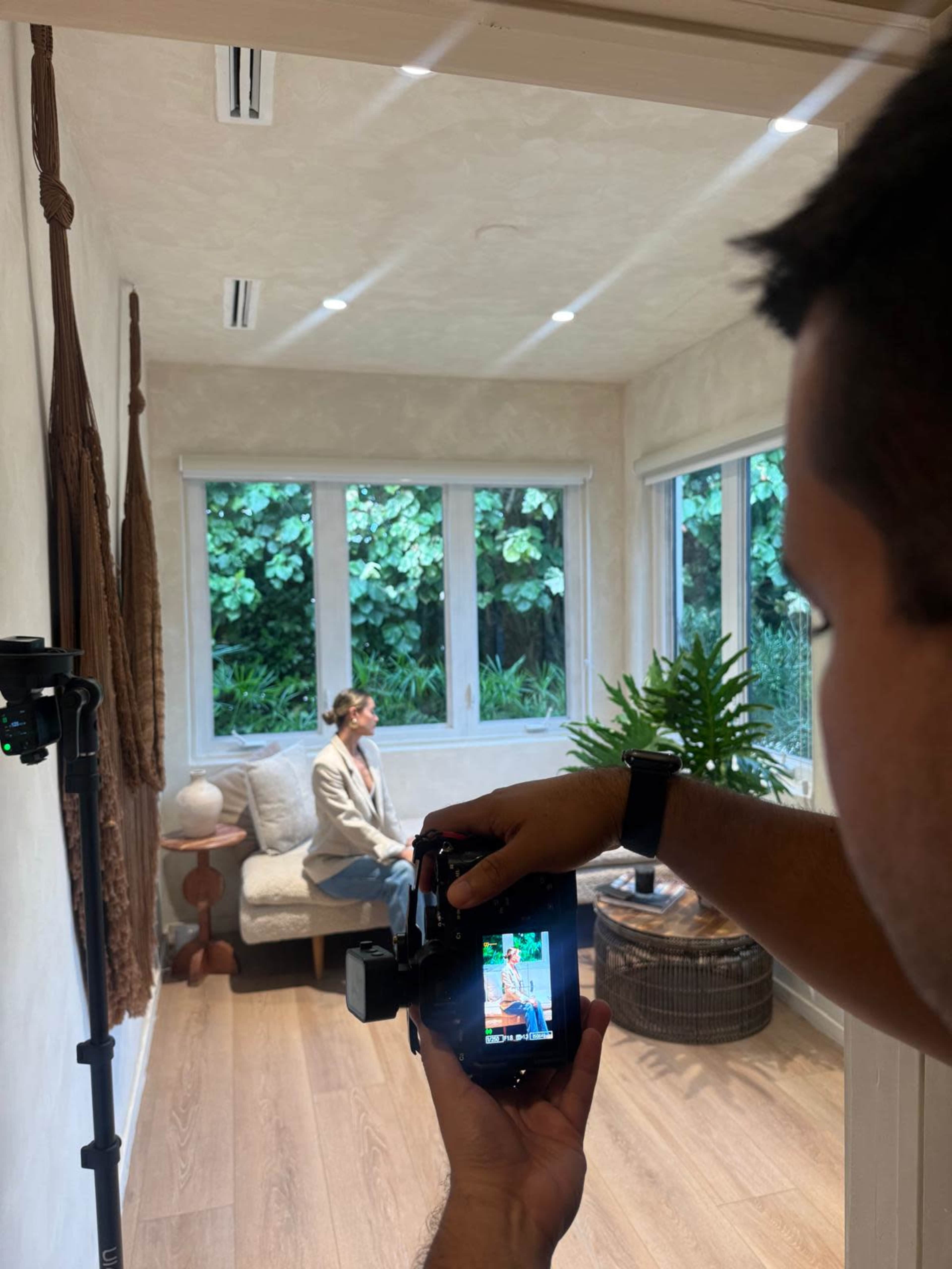 A person is taking a photograph of a seated individual in a room filled with natural light and greenery outside.