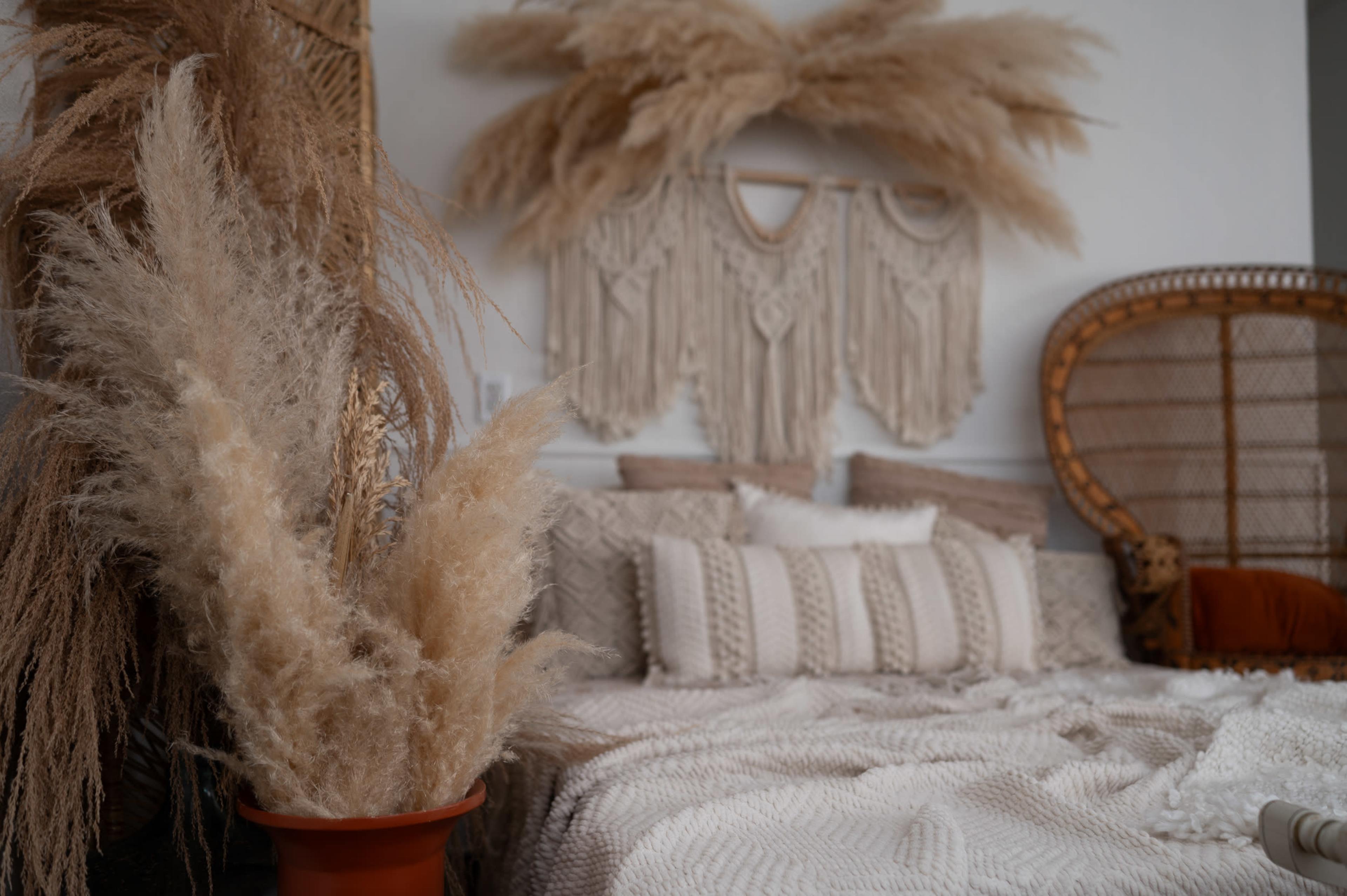 A cozy bedroom features a woven wall hanging, a bed adorned with knitted blankets and pillows, and a decorative chair alongside a large pampas grass arrangement in a terracotta pot.