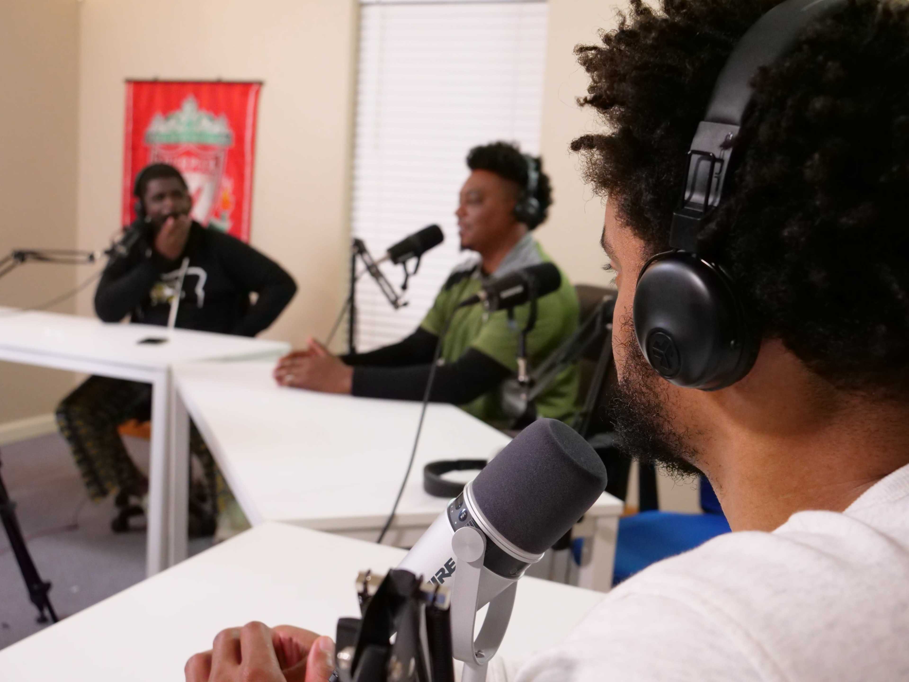 Three individuals participate in a podcast recording, seated at a table with microphones in front of them.