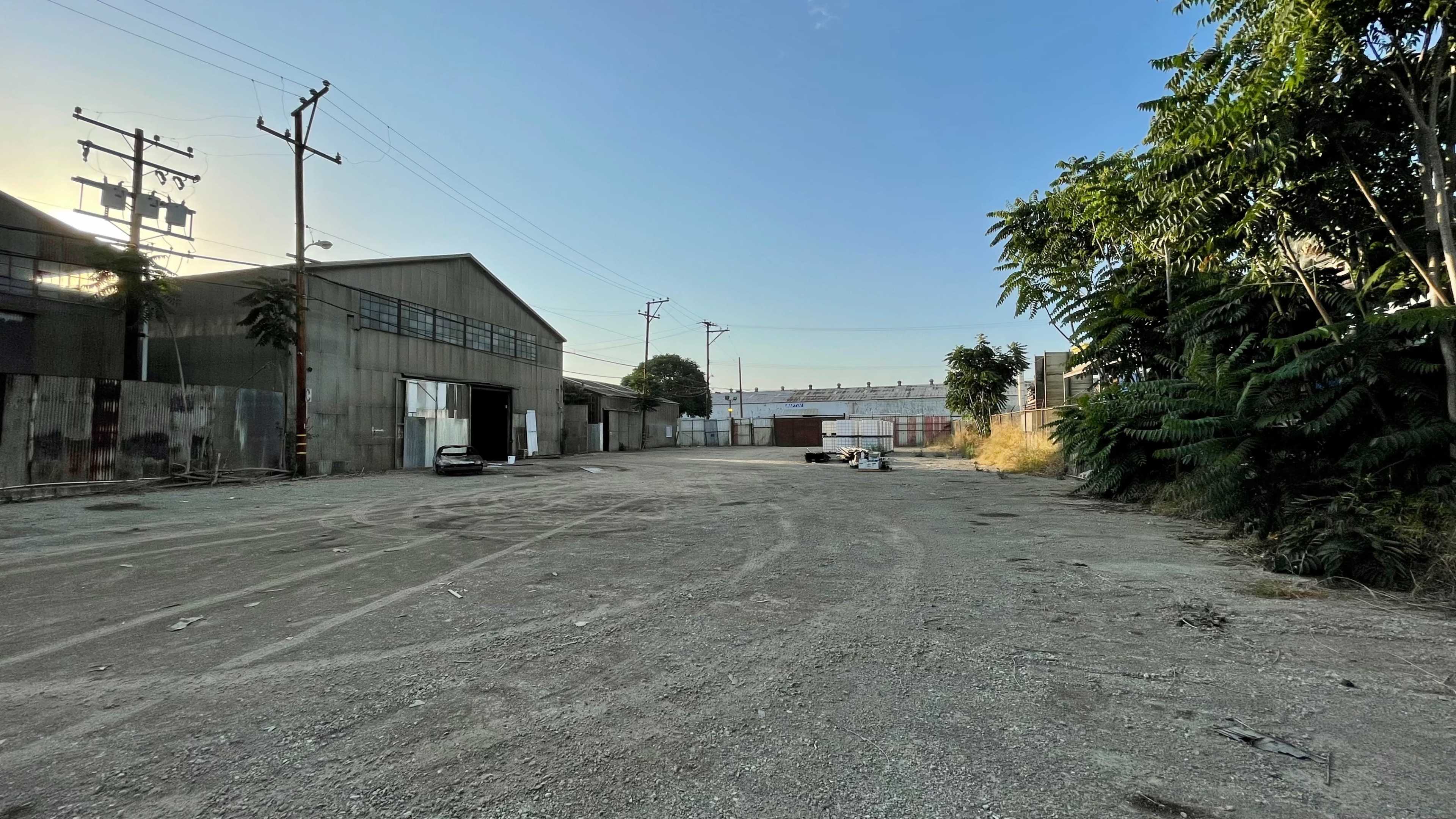 A vacant industrial area with gravel ground, surrounded by warehouses and overgrown foliage.
