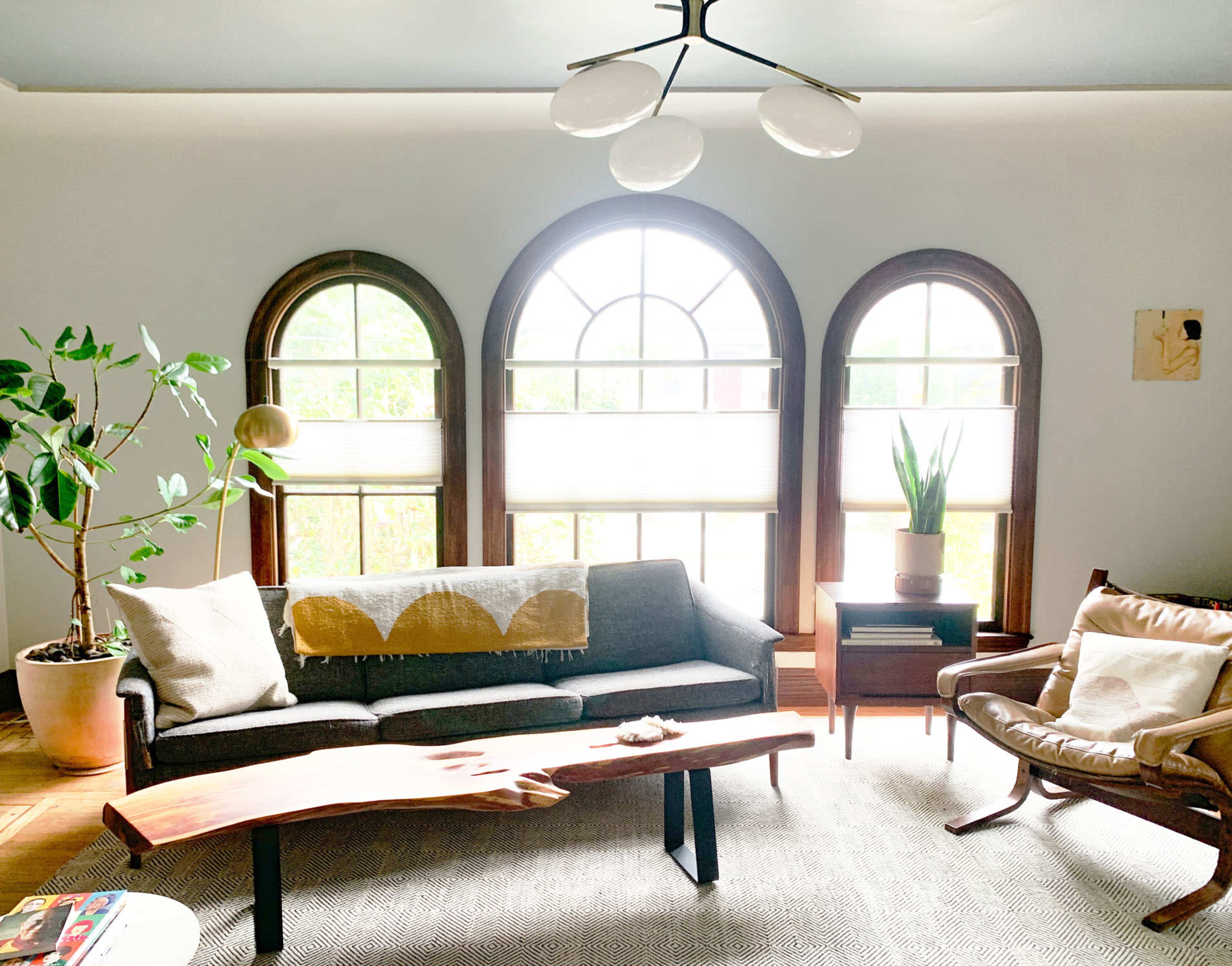 A living room features large arched windows, a gray sofa with a decorative throw, a wooden coffee table, and potted plants.