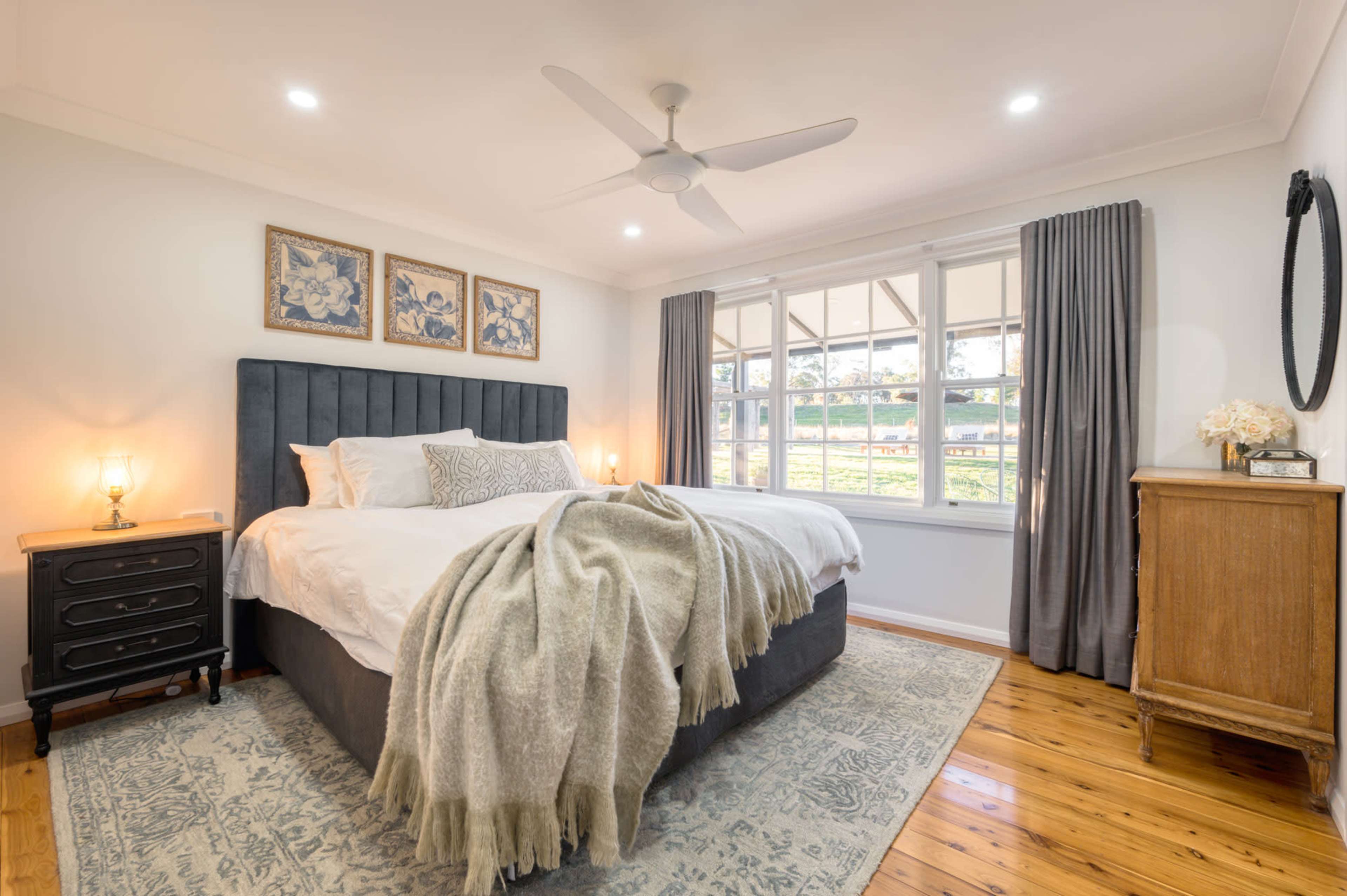 A cozy bedroom featuring a large bed with a gray upholstered headboard, white bedding, and a patterned area rug, accompanied by wooden furniture and large windows allowing natural light.