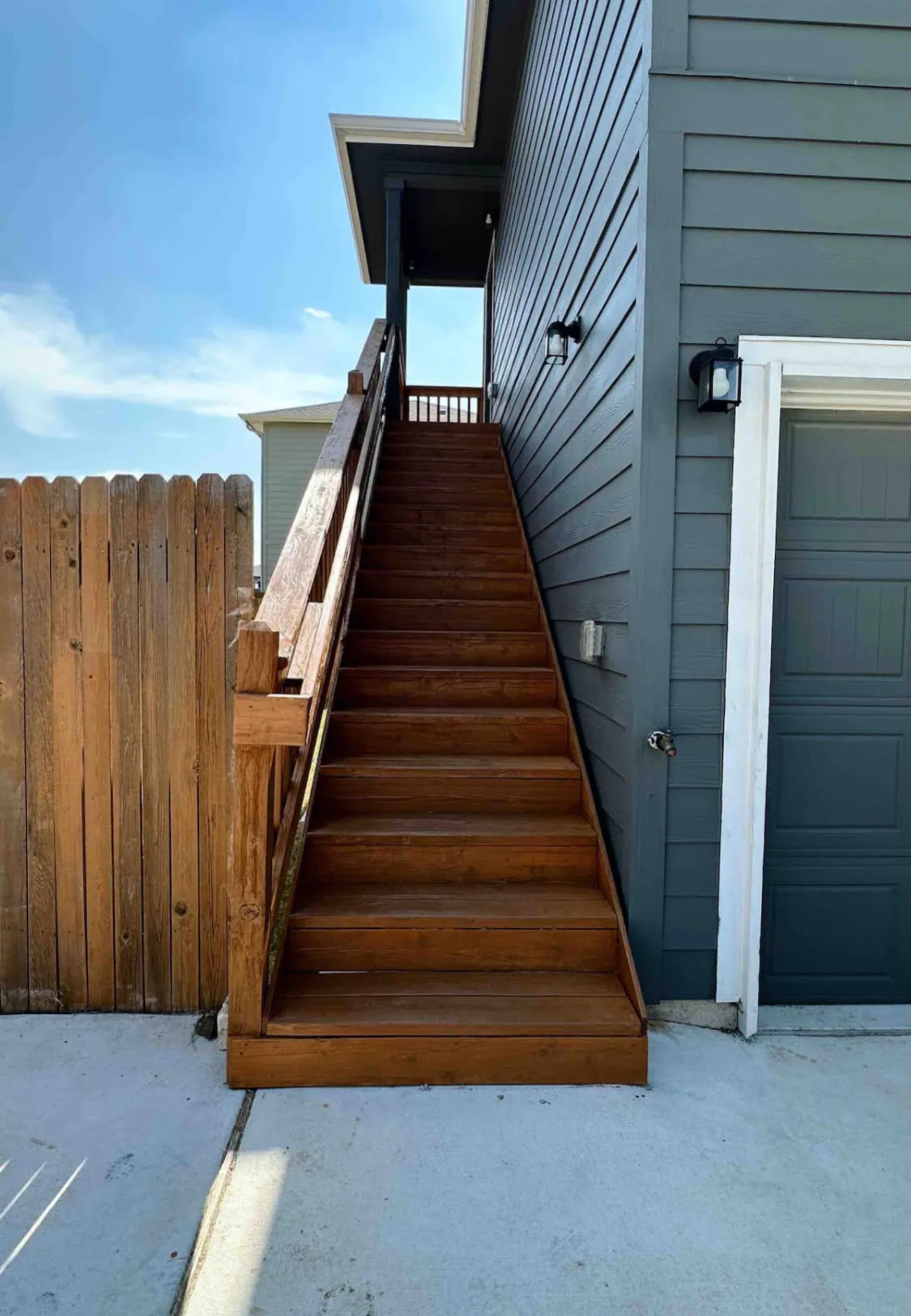 A set of wooden stairs leads up to a door on the side of a gray house, next to a wooden fence.