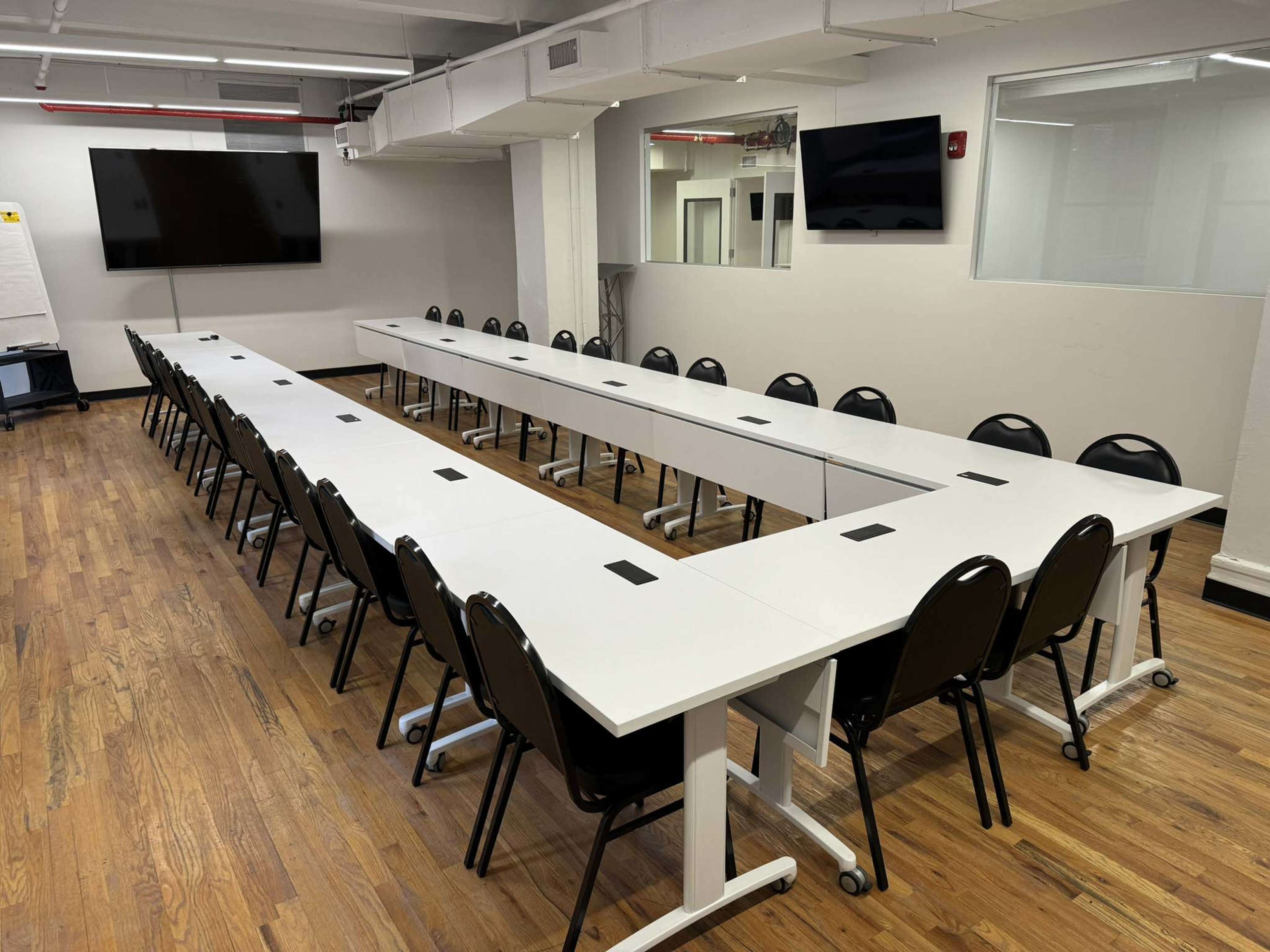 A rectangular conference room features long white tables arranged in a U-shape with black chairs along the sides.