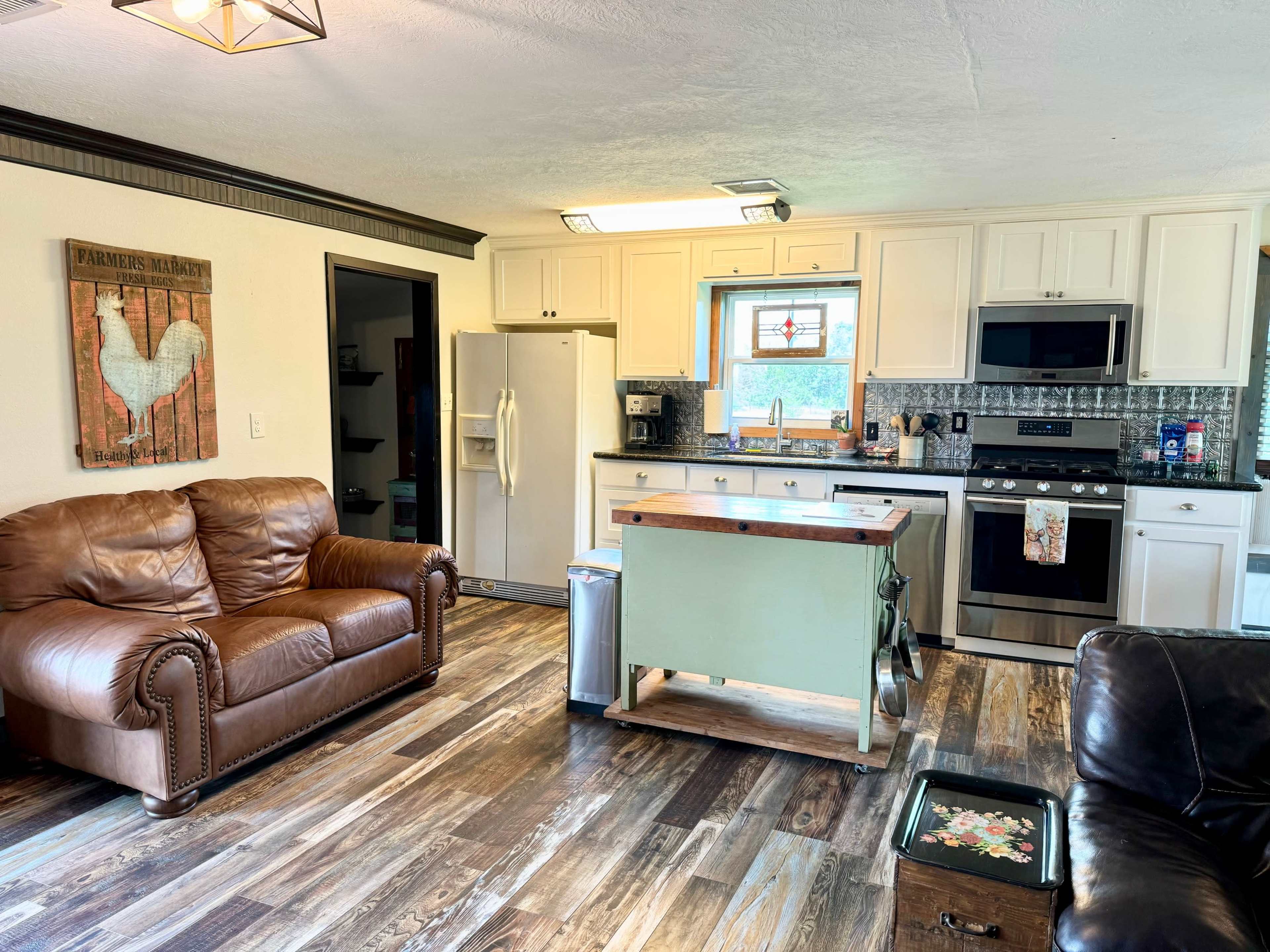 The image shows a cozy kitchen connected to a living area, featuring wooden cabinets, a central island, and two leather sofas.