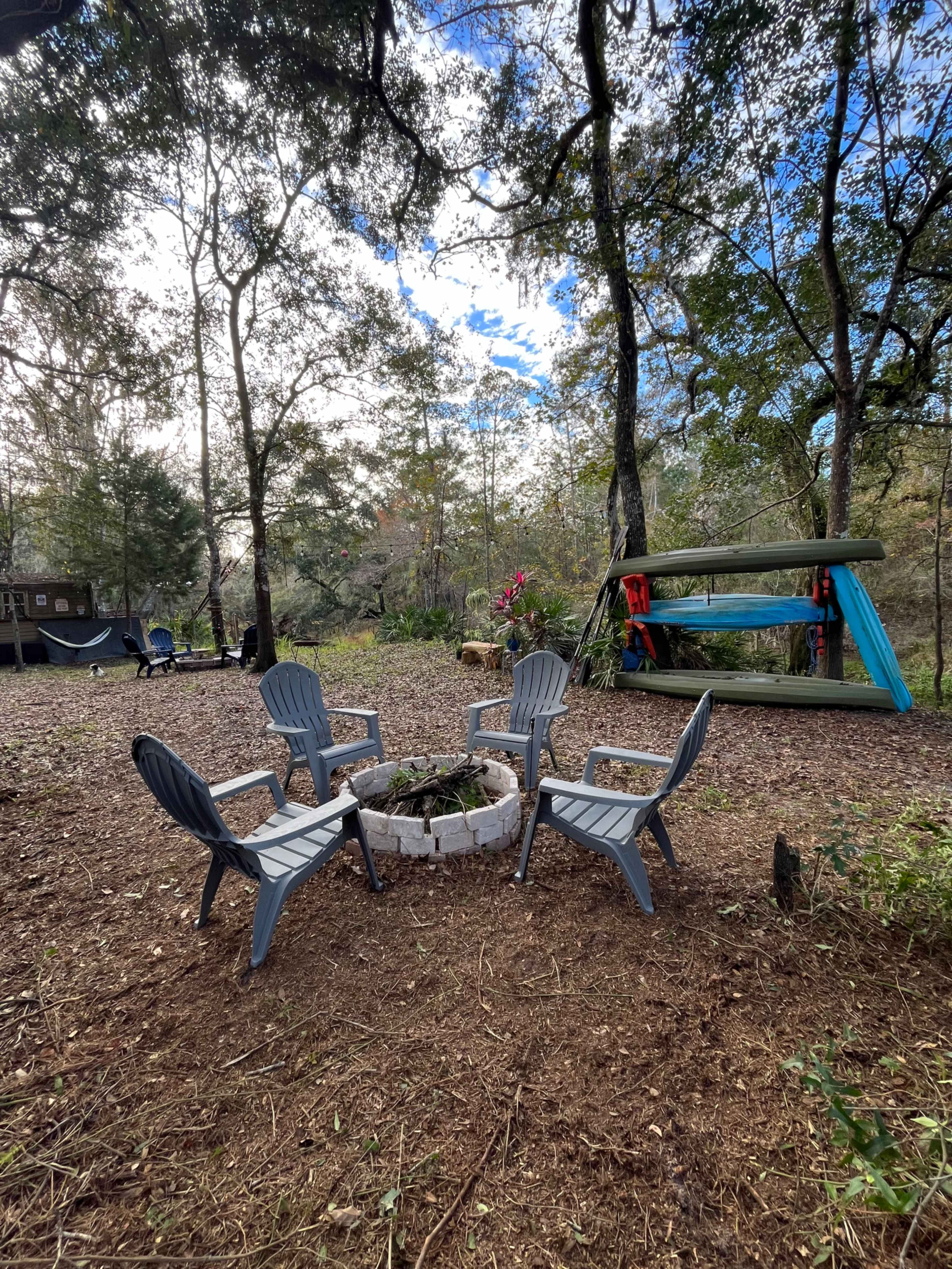 A fire pit surrounded by four gray chairs in a wooded area with a hammock and a colorful wooden structure in the background.