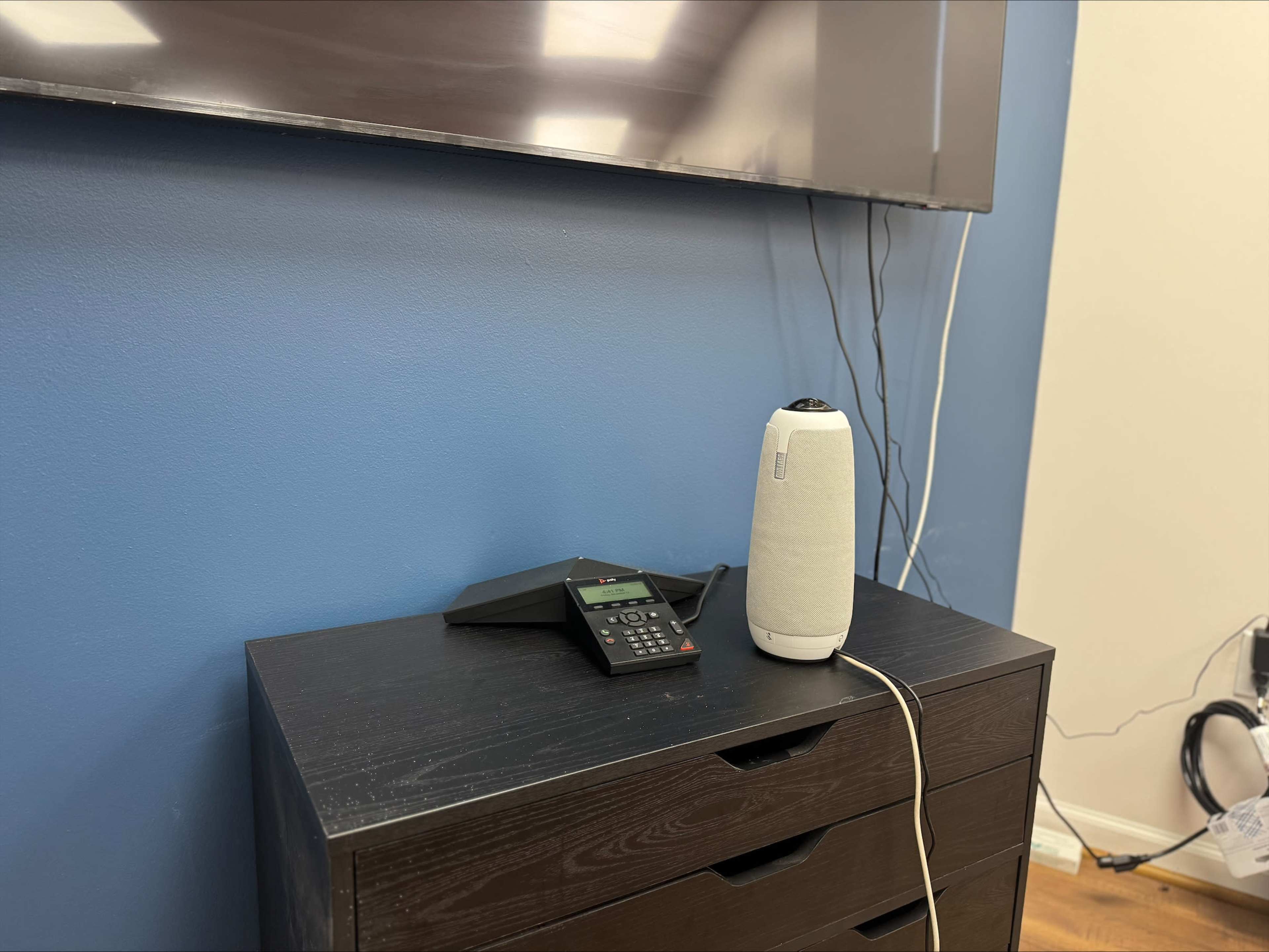 A black dresser holds a speaker and a phone, positioned against a blue wall beneath a television screen.