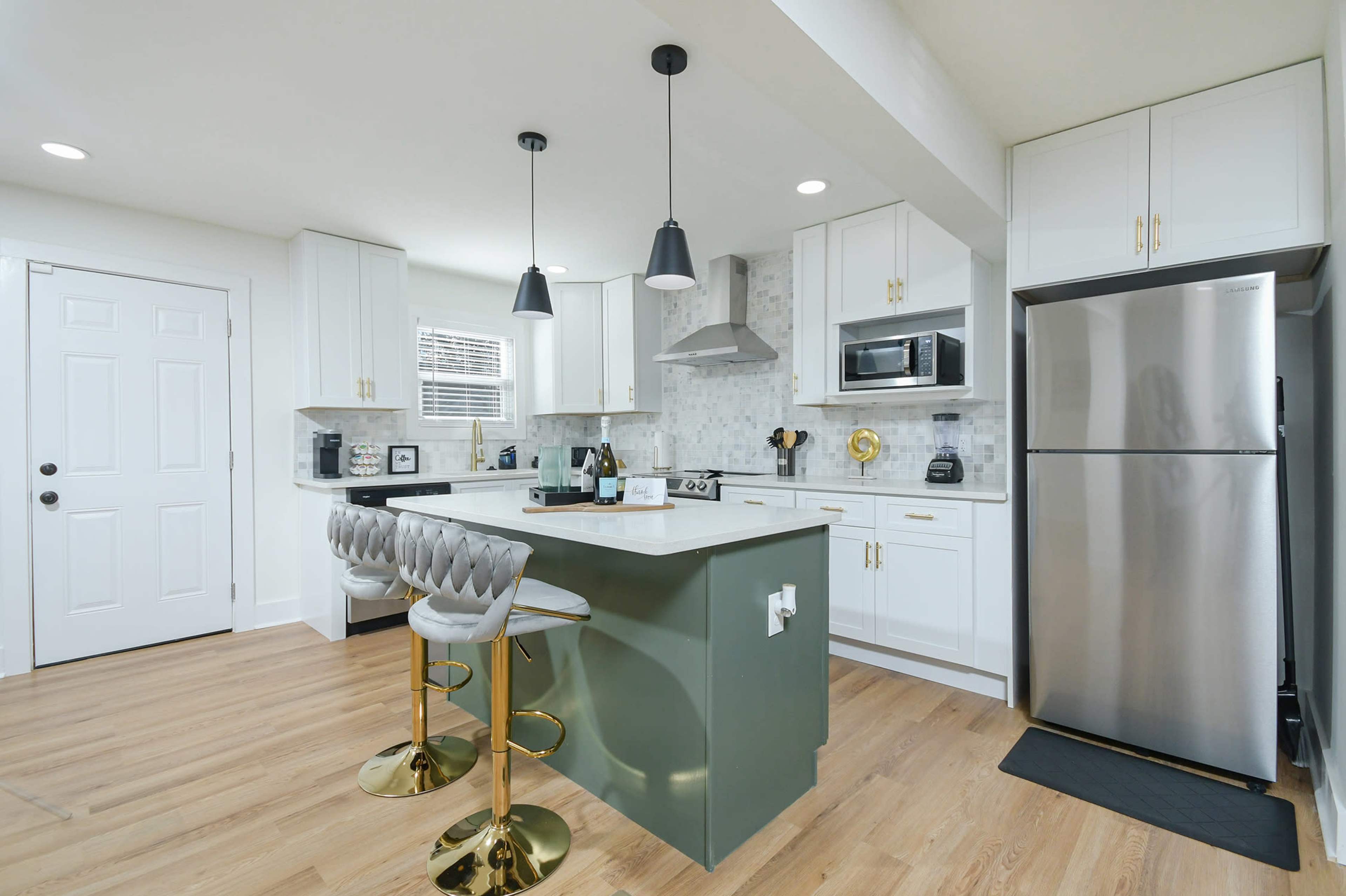 The image shows a modern kitchen with white cabinets, sleek stainless steel appliances, a green island, and three gold-accented bar stools.
