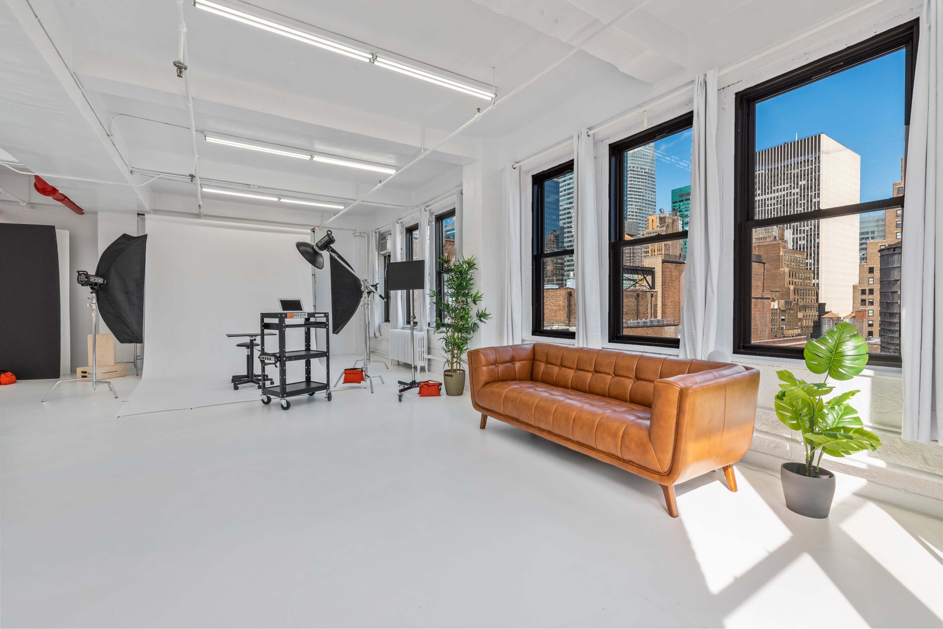 A bright, modern studio space featuring a brown leather couch, large windows with city views, and a setup for photography with a backdrop and lighting equipment.