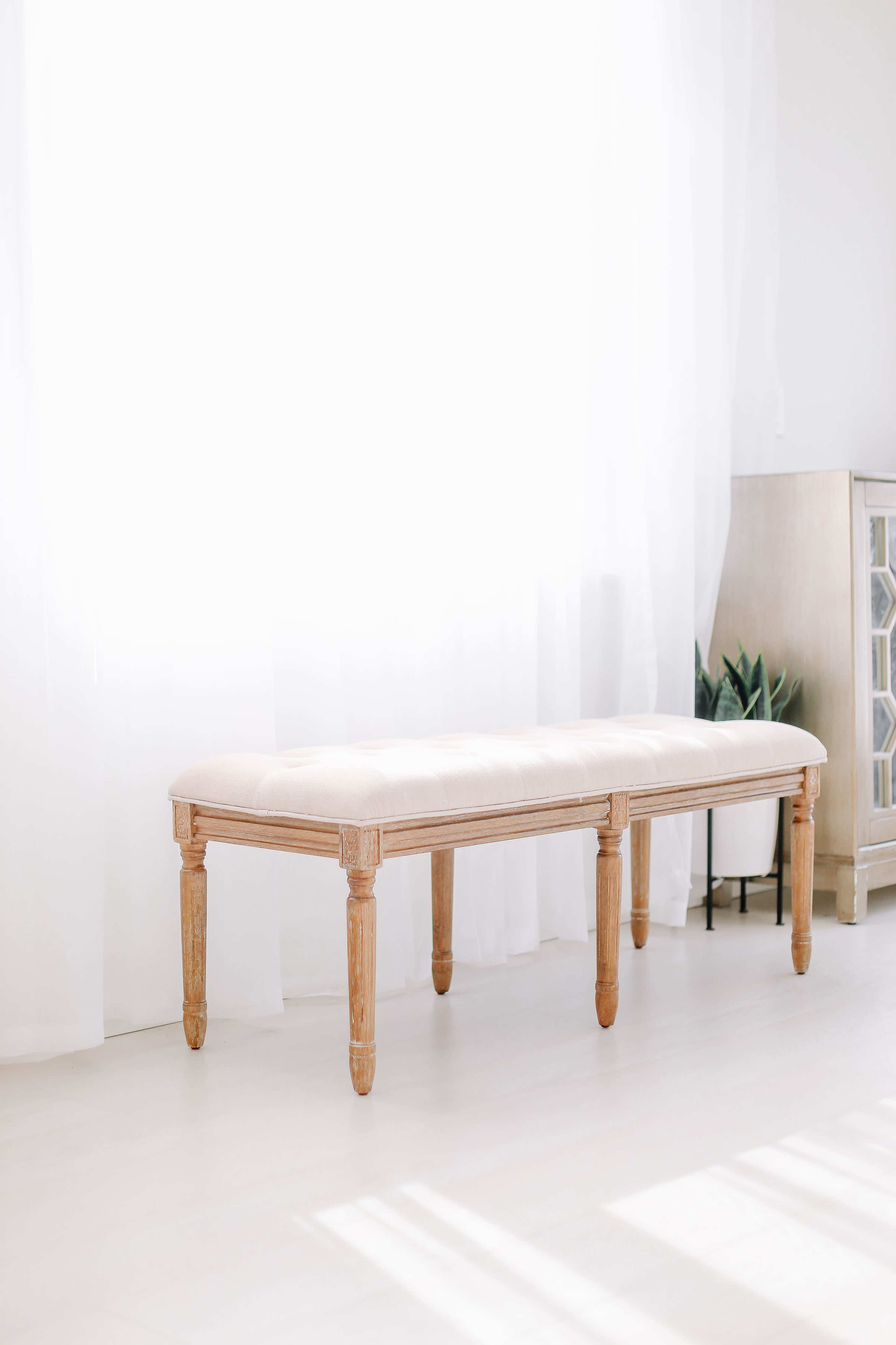 A wooden bench with a padded seat is positioned in a brightly lit room with sheer curtains.
