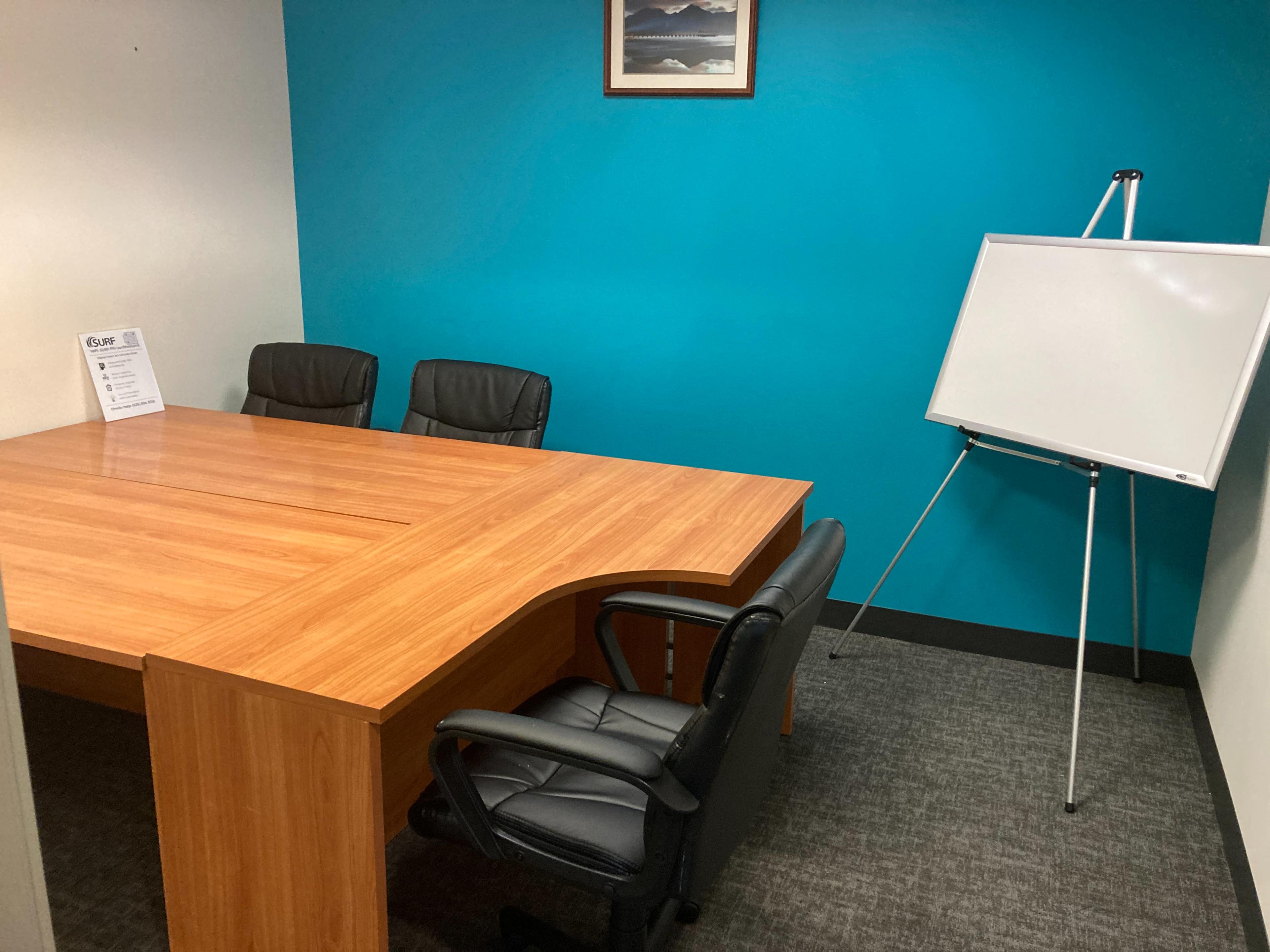 A small meeting room features a wooden table, three black chairs, and a whiteboard against a blue wall.