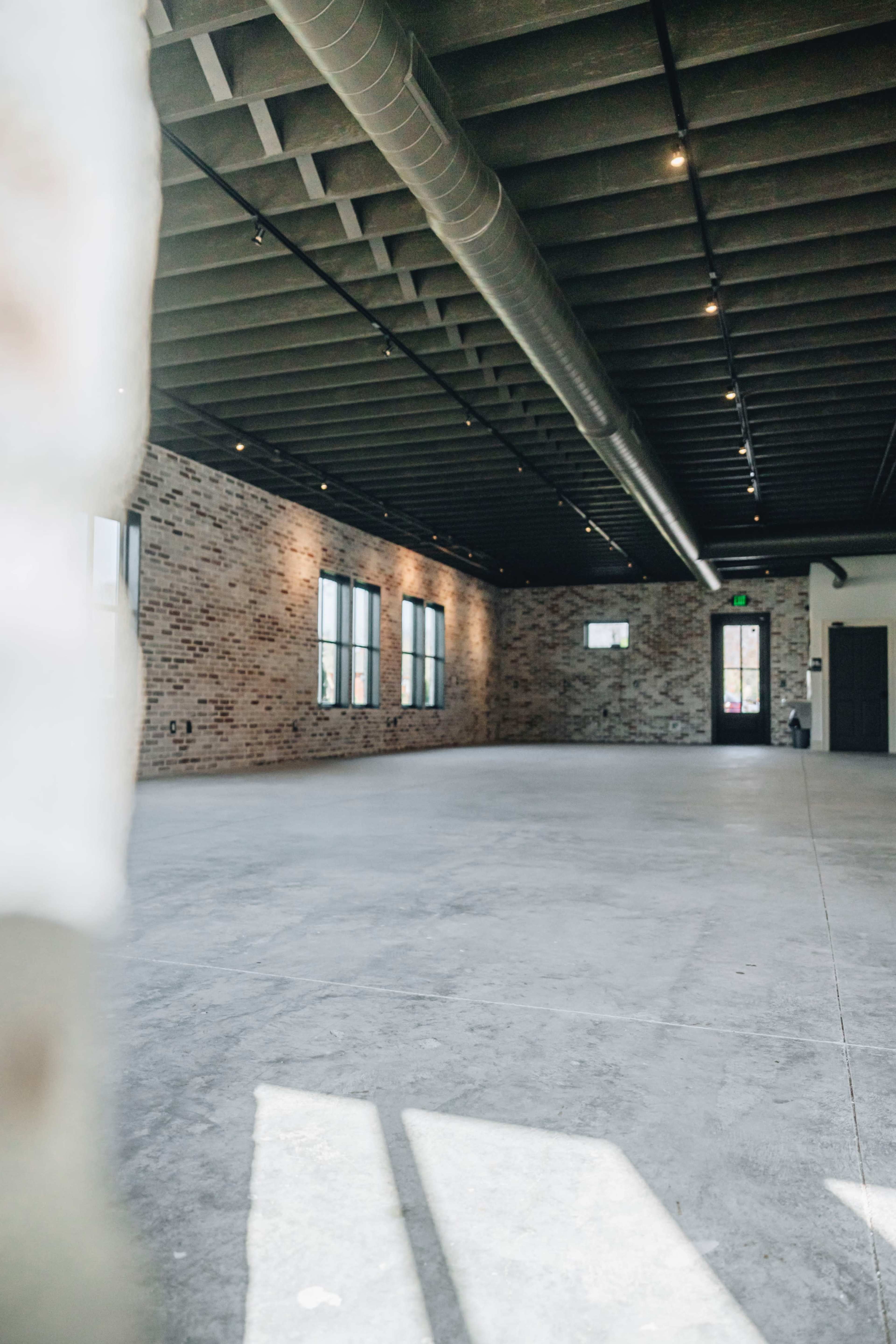 A spacious, industrial-style room with exposed brick walls, large windows, and a concrete floor.