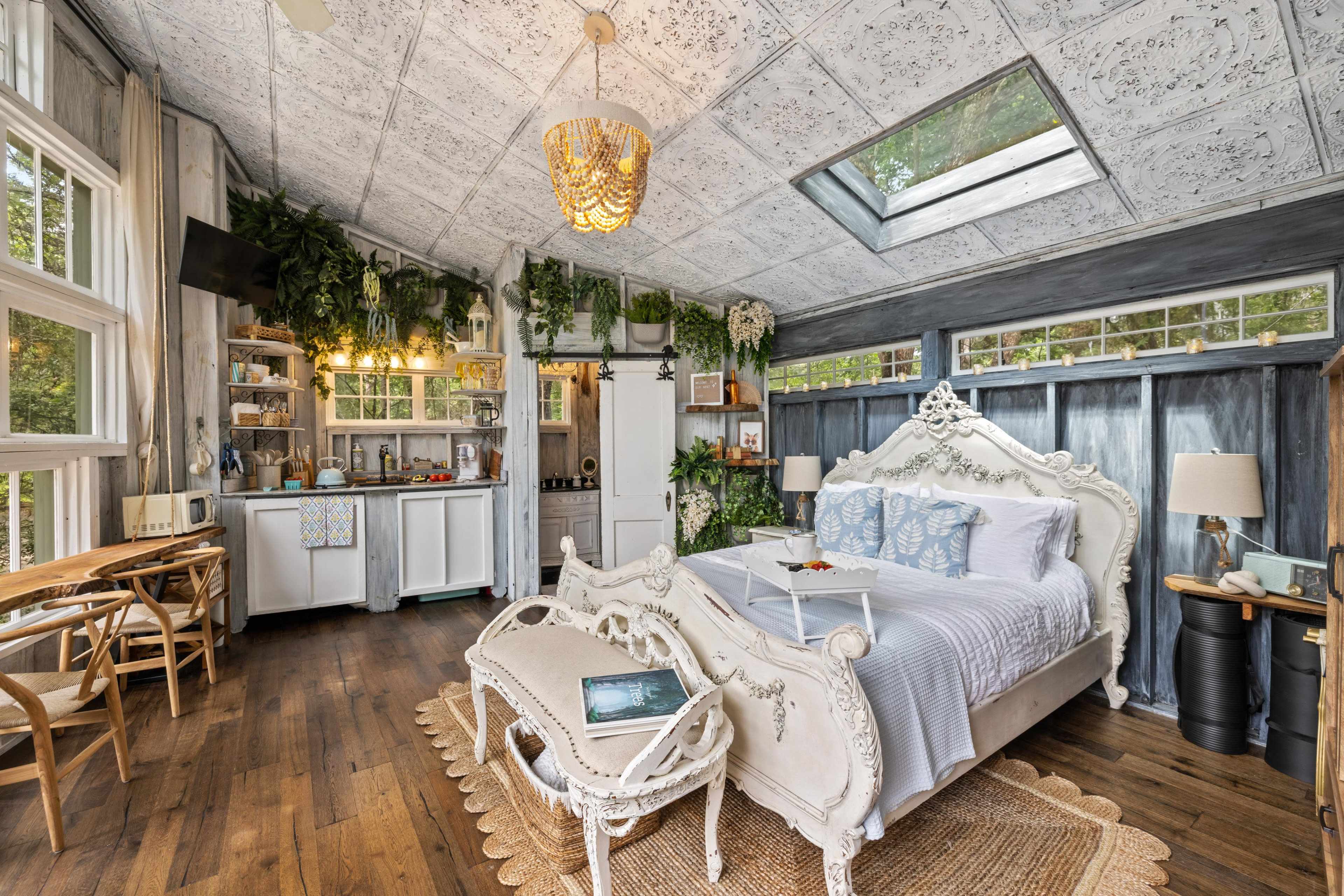 A cozy bedroom with a large bed, a small kitchenette area, and several plants decorating the walls and windows.