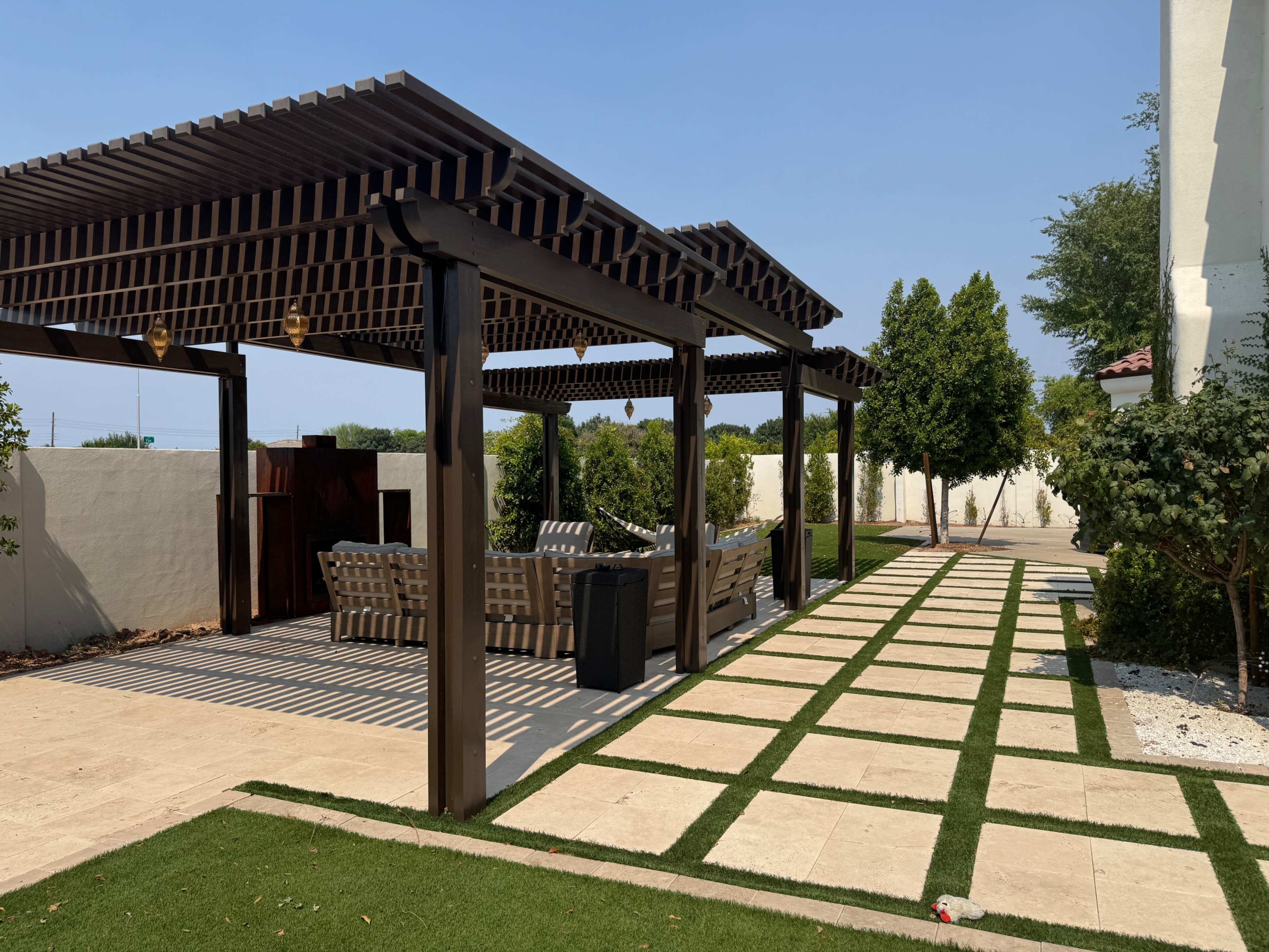 The image shows a landscaped outdoor area featuring a wooden pergola, a seating arrangement, and a pathway lined with grass and stone pavers.