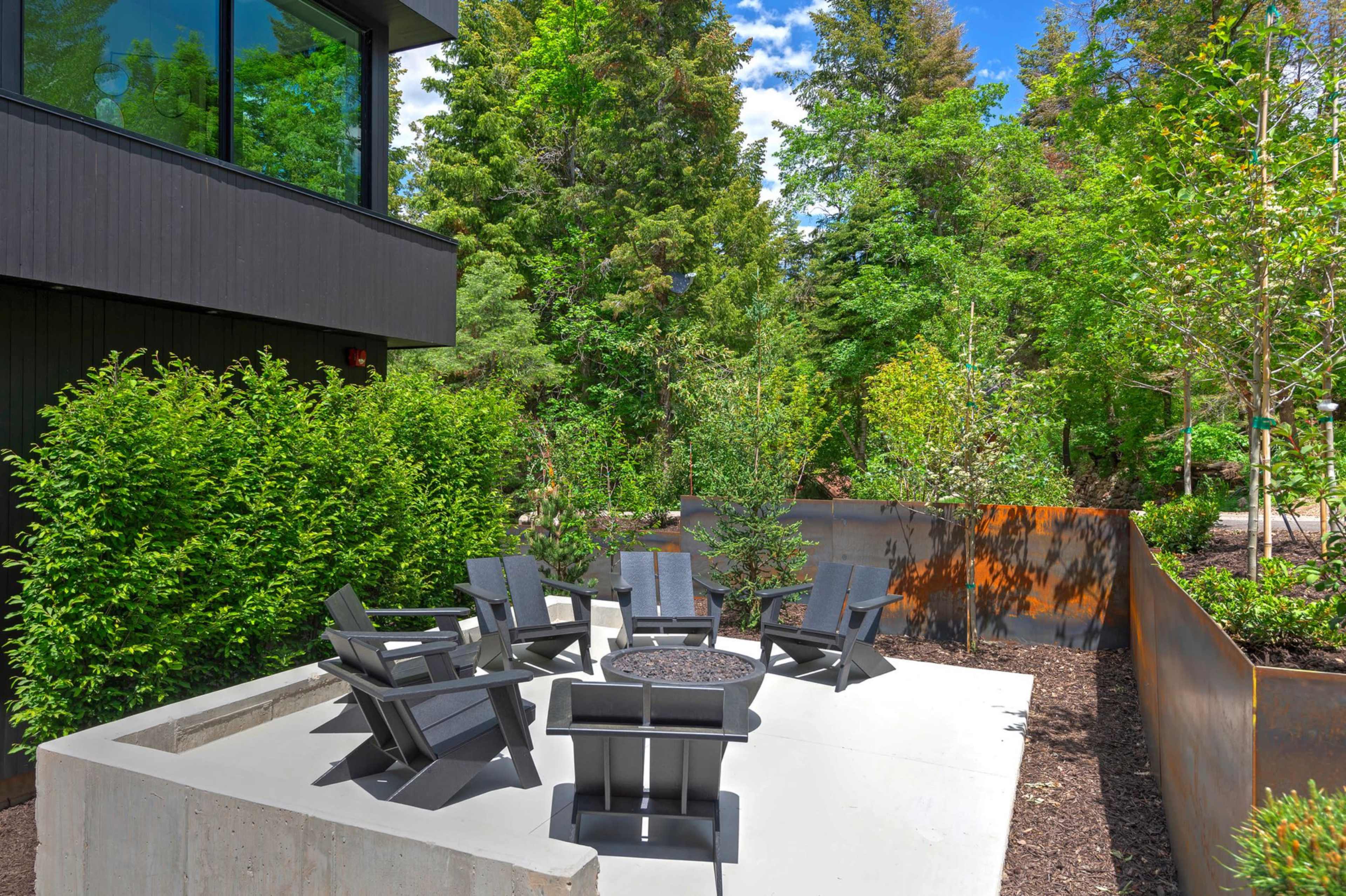 A concrete patio with five black chairs and a central fire pit is surrounded by lush greenery and trees.