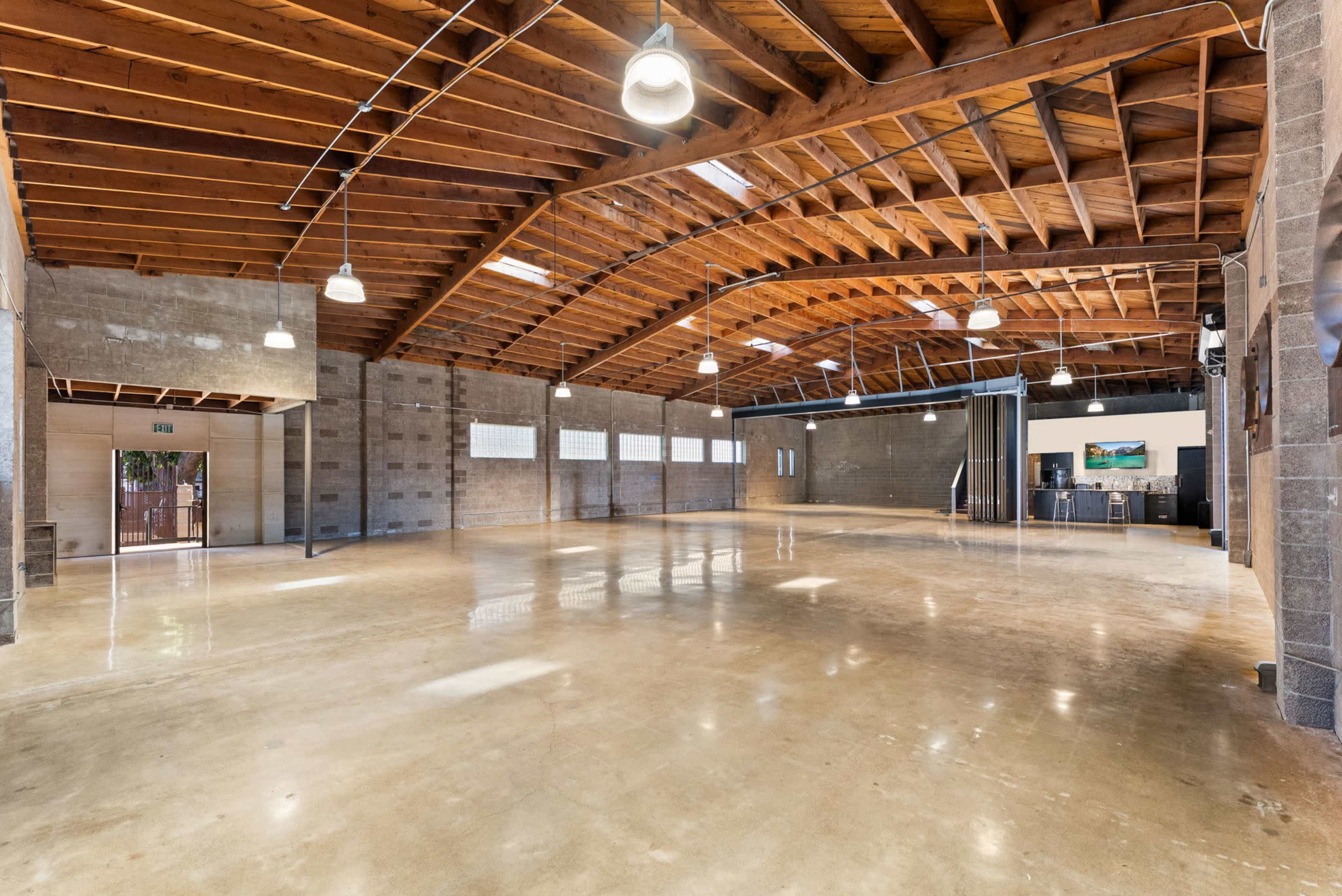 The image shows a spacious, industrial-style room with a high wooden-beam ceiling, polished concrete floors, and large windows allowing natural light to enter.