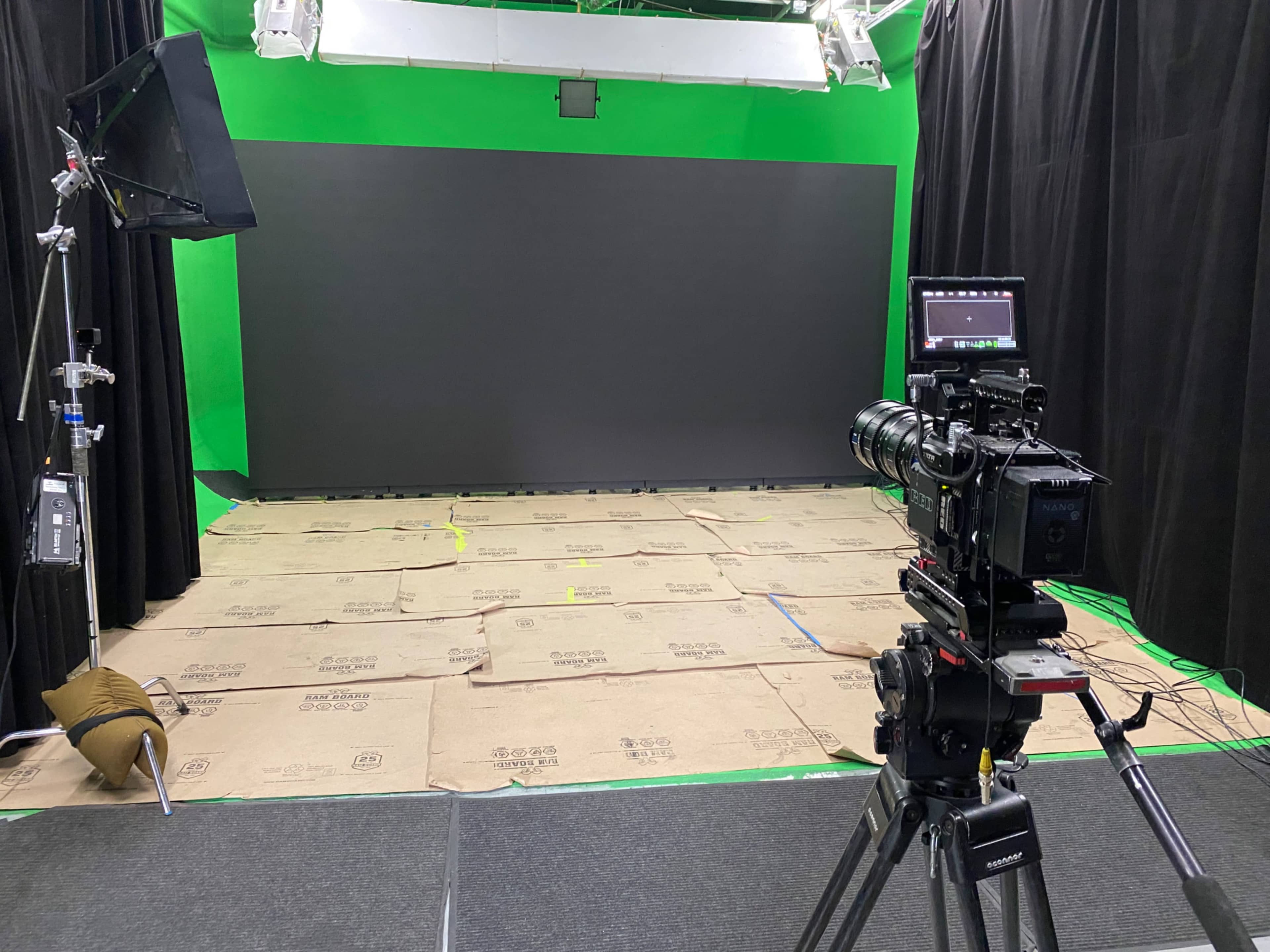 The image shows a film or photo studio setup with a green screen backdrop, a camera on a tripod, and cardboard covering the floor.