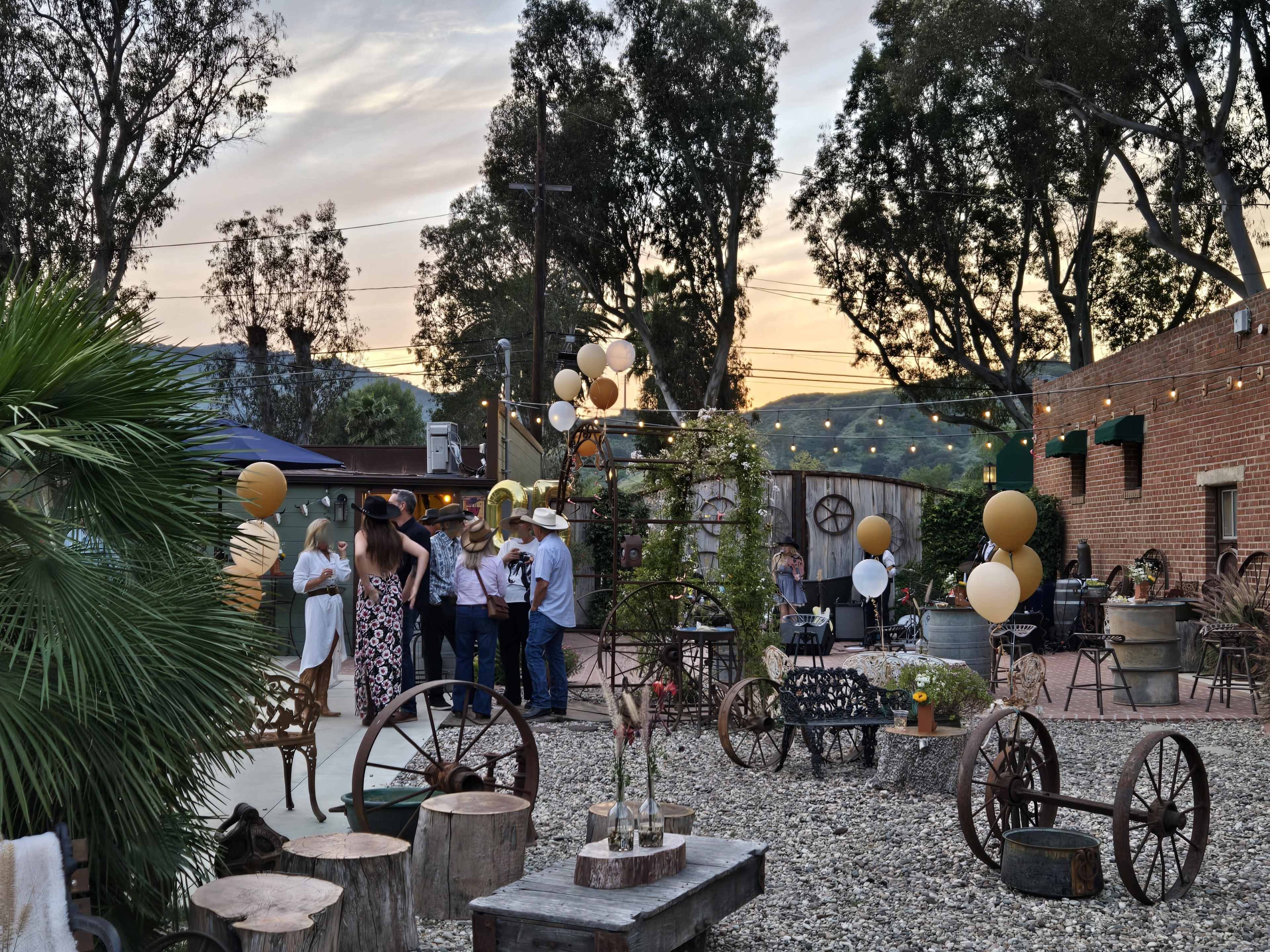The scene features a rustic outdoor gathering with guests mingling among decorated tables and trees, illuminated by string lights under a colorful sunset.