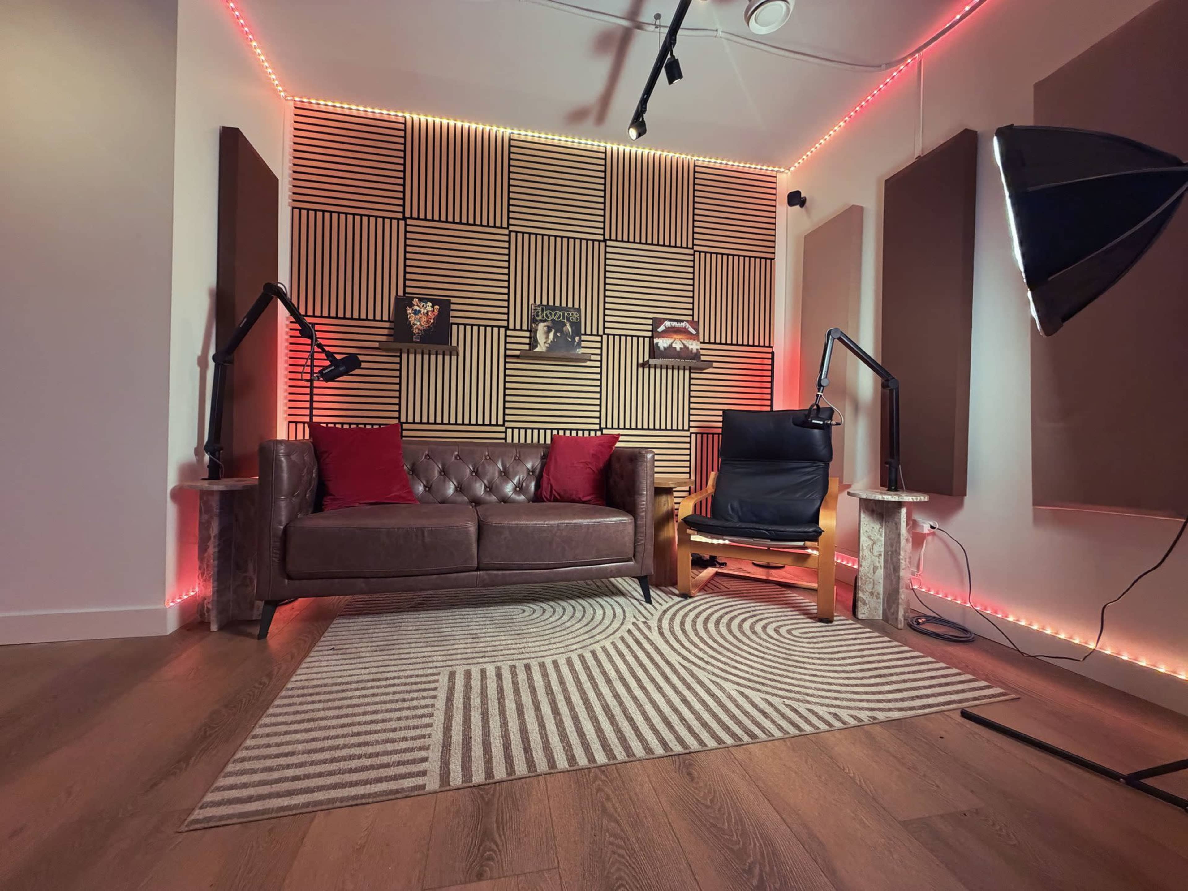 The image shows a cozy studio space featuring a brown leather couch and a black armchair, with wooden paneling on the walls and soft lighting.
