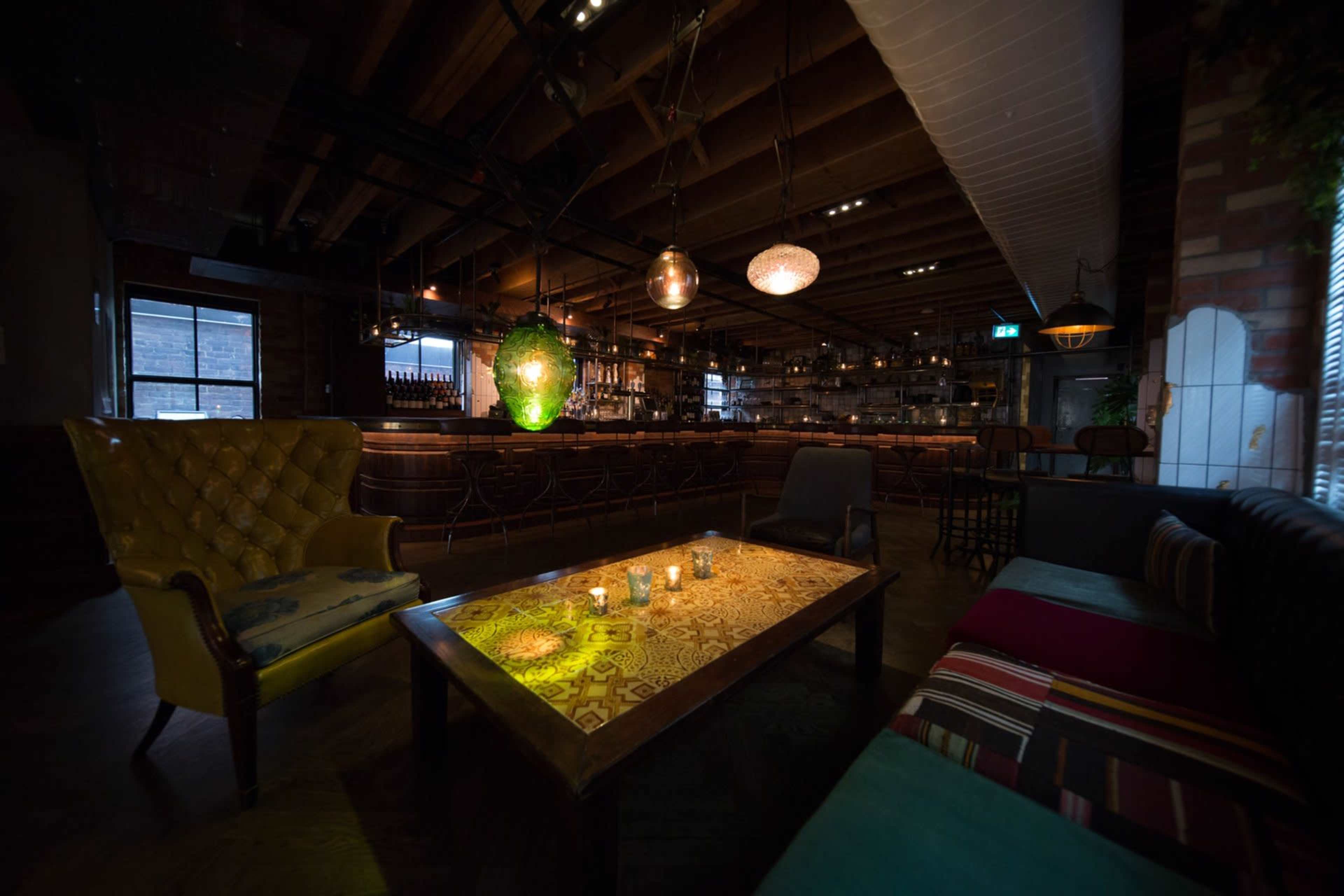 The image shows a dimly lit restaurant interior featuring colorful seating arrangements and a wooden table with candles on it.