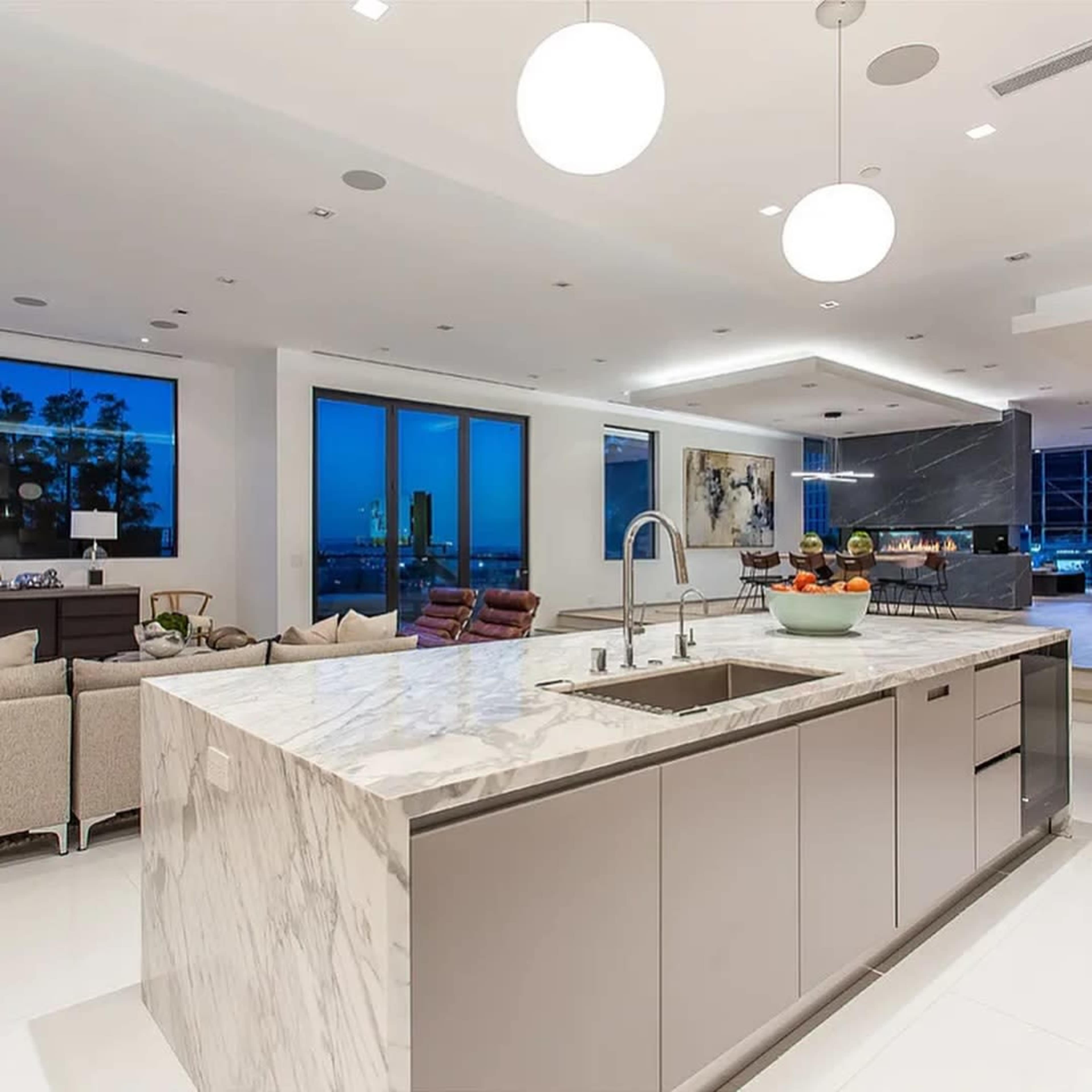 The image shows a modern kitchen with a large marble island, stainless steel fixtures, and floor-to-ceiling windows revealing a city view at dusk.