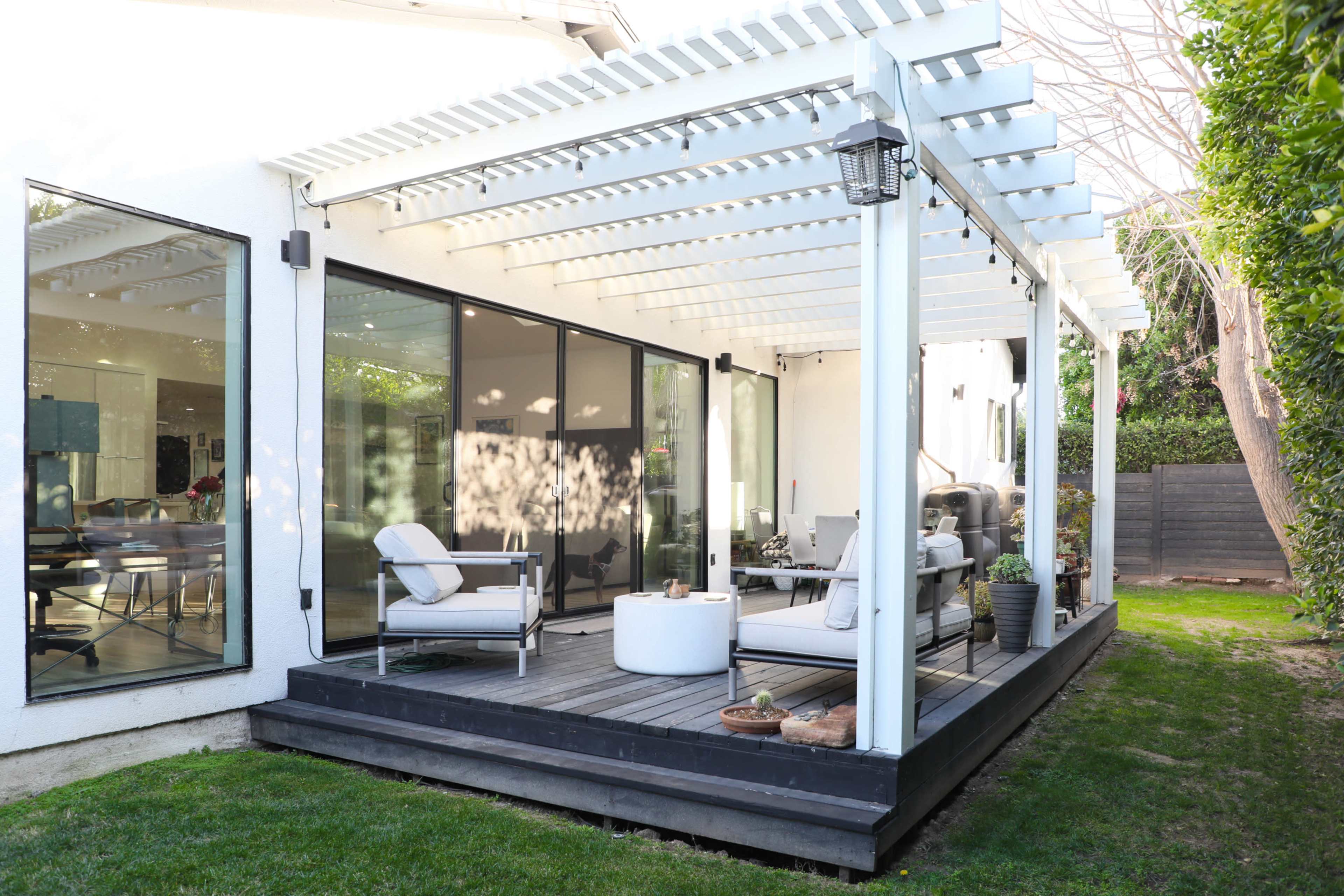 A modern patio with a covered seating area, featuring a white pergola and a mix of furniture on a wooden deck surrounded by green grass.