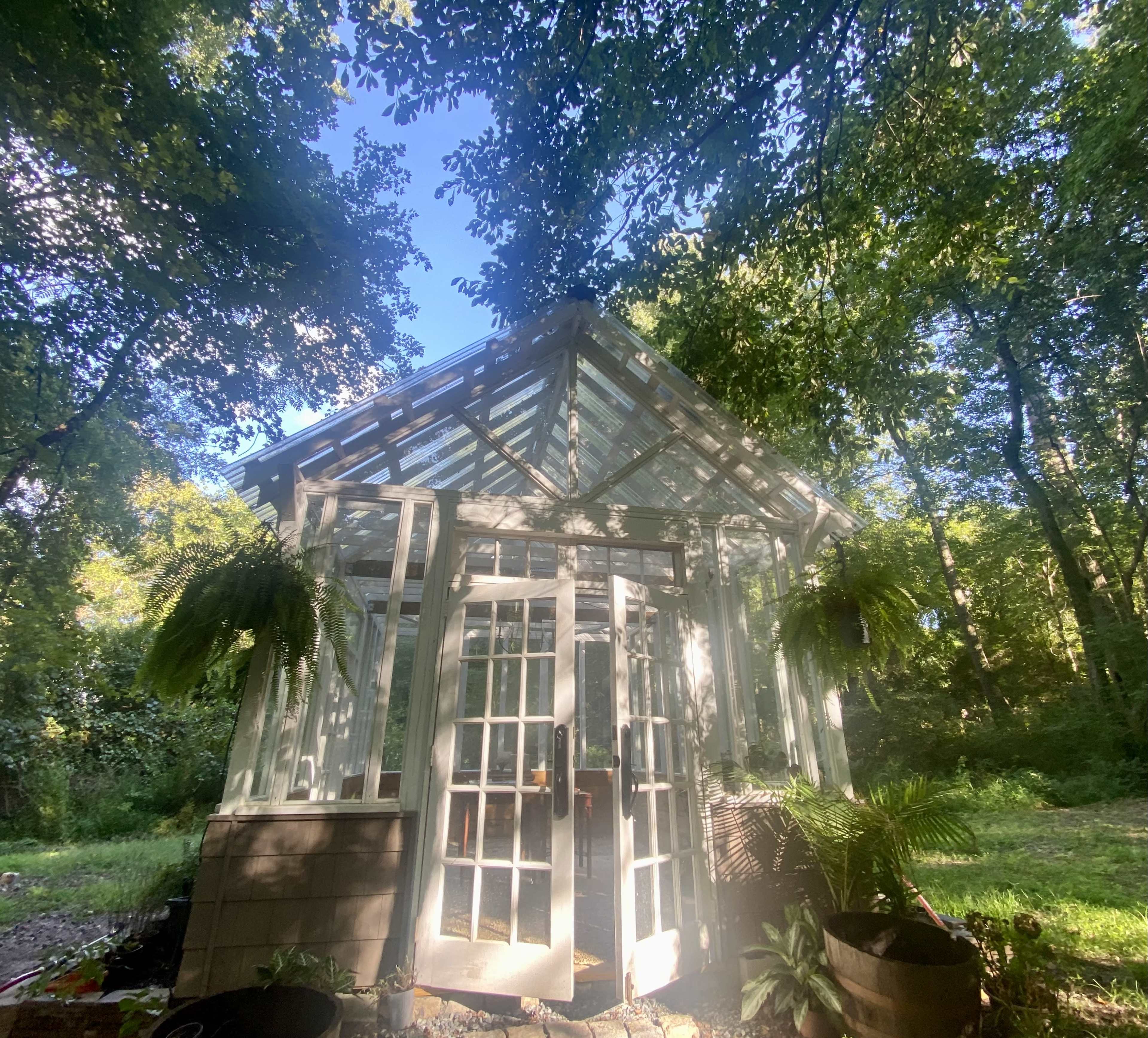 A glass greenhouse with double doors is surrounded by trees and greenery.
