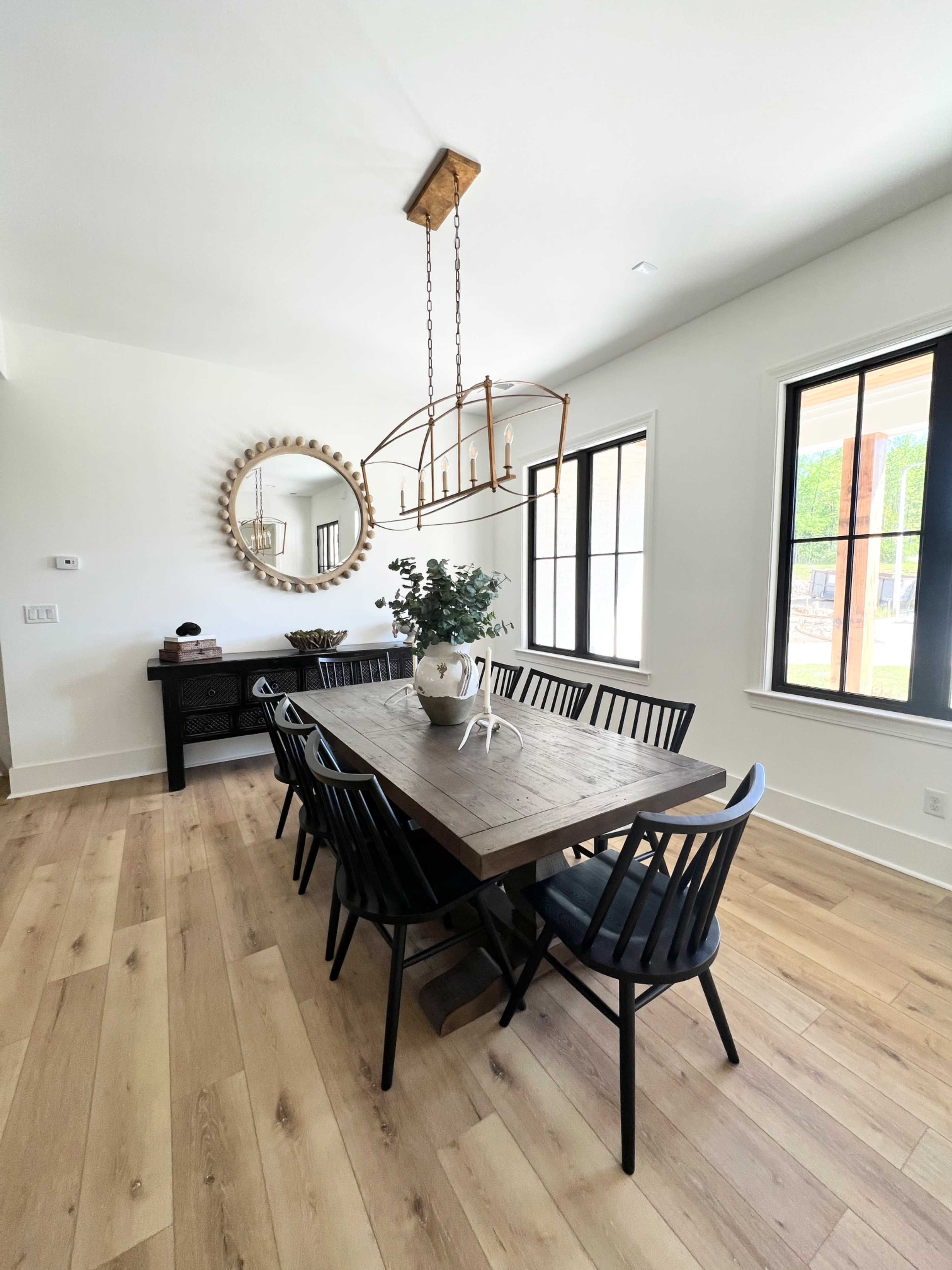 A large wooden dining table is surrounded by black chairs in a bright room with a round mirror and a wooden light fixture.
