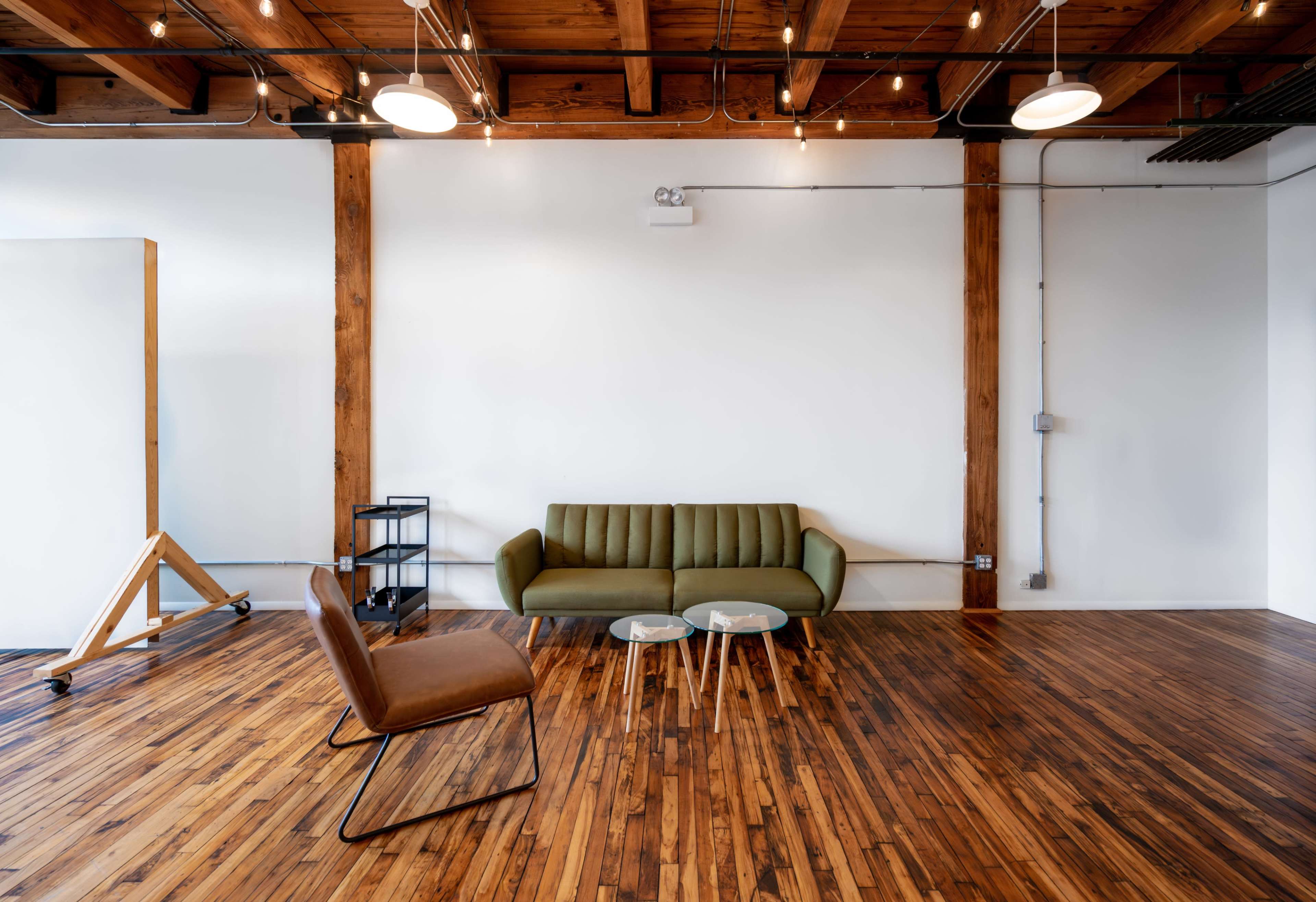 A minimalist living space featuring a green sofa, a brown armchair, and two glass tables on wooden flooring with exposed wooden beams above.