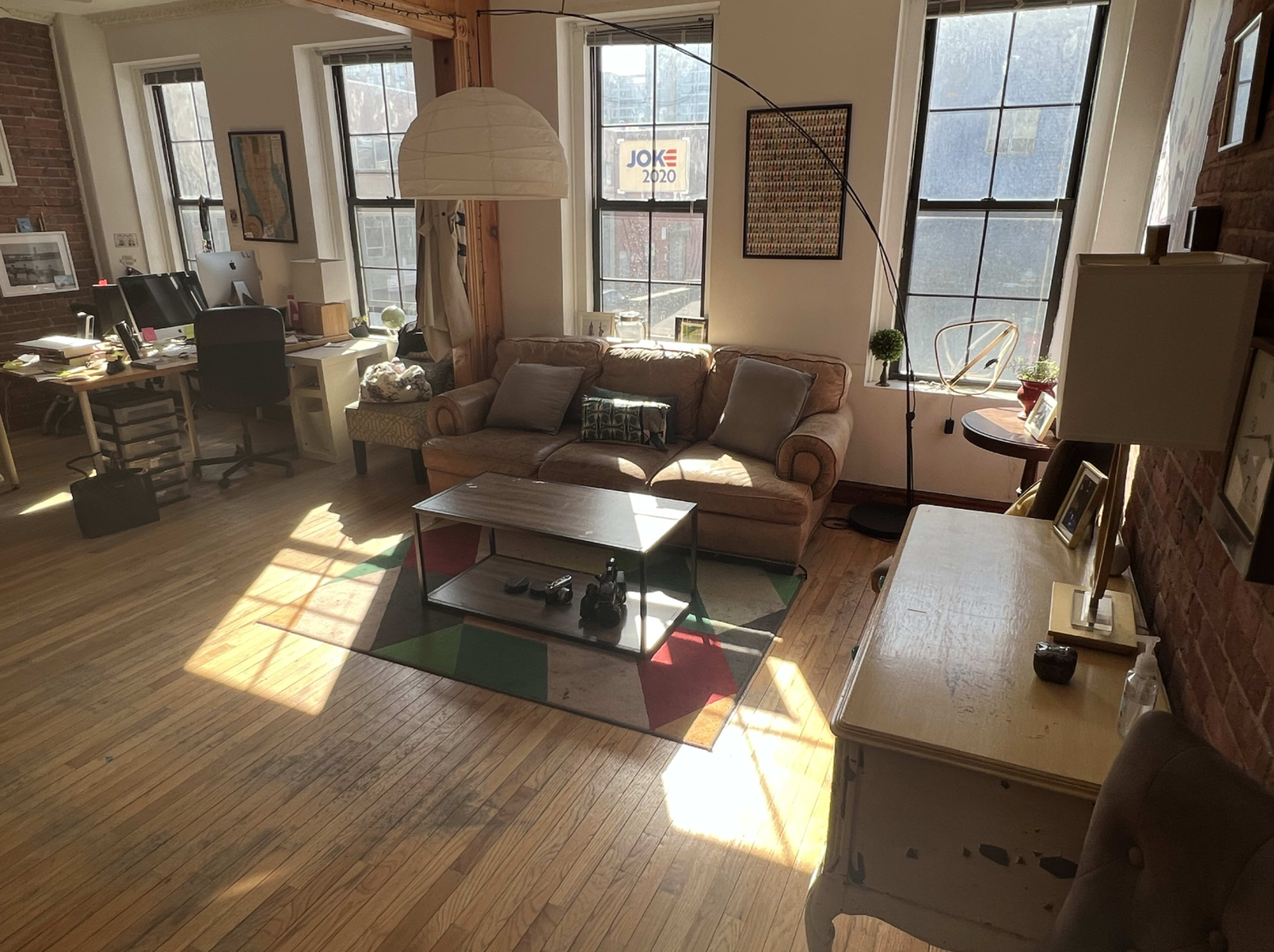 A sunlit living room with a sofa, a coffee table, and a desk area, featuring large windows and wooden flooring.