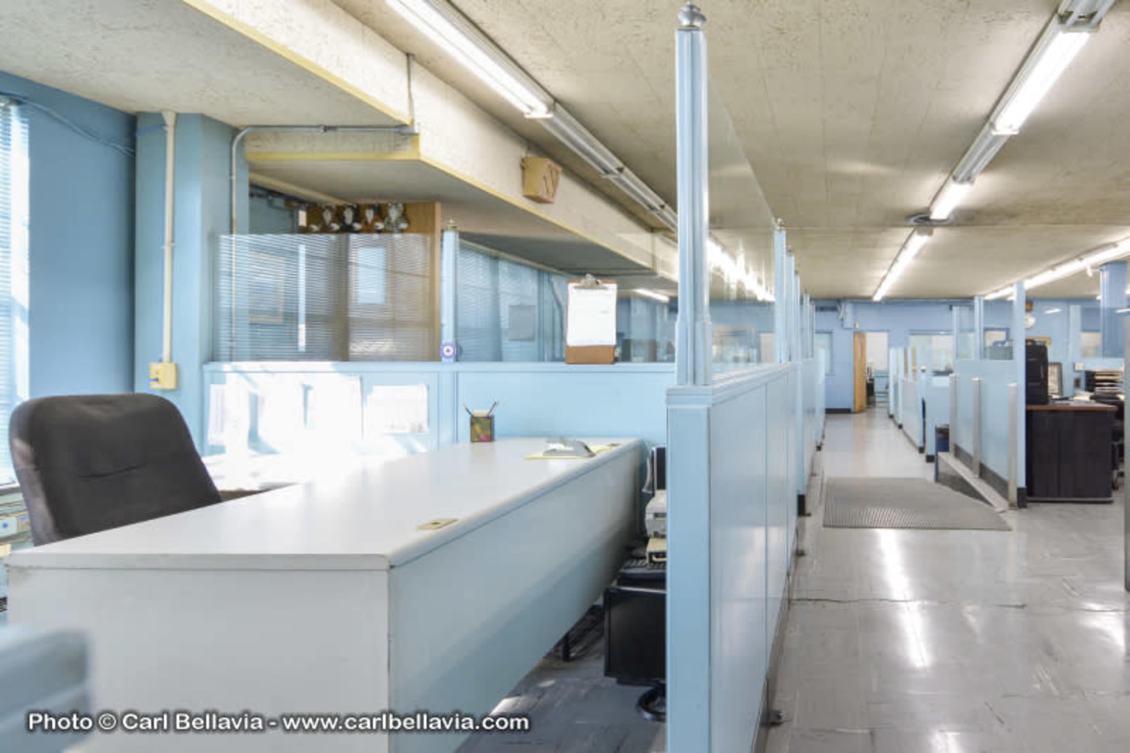 A spacious office layout with desks and partitions, illuminated by overhead lights.
