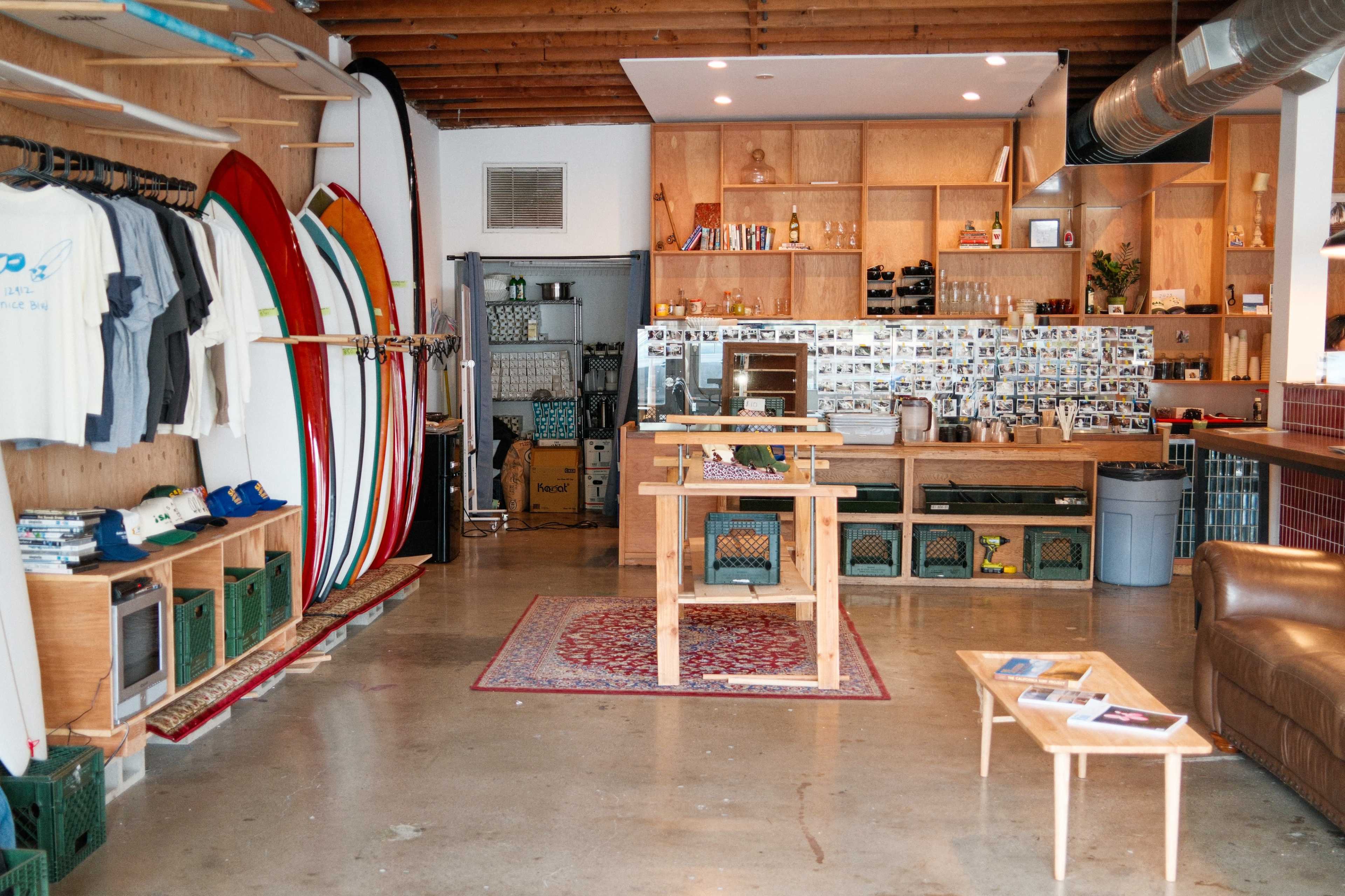 The image shows an interior of a surf shop with surfboards displayed on one side, clothing racks, a wooden table in the center, and a seating area with a couch.