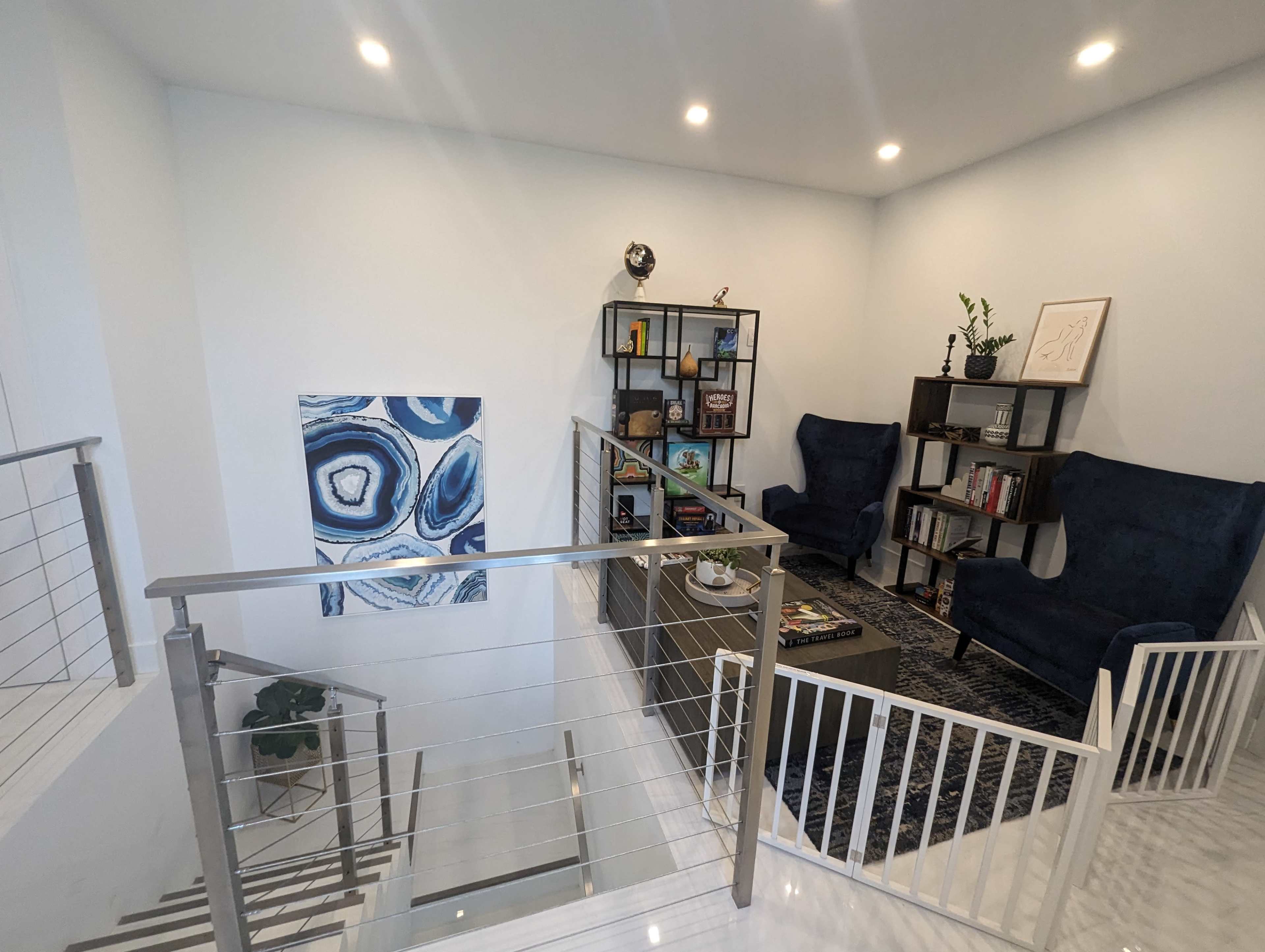 A modern second-floor landing with two blue chairs, a small table, a bookshelf, and a framed abstract painting on the wall.