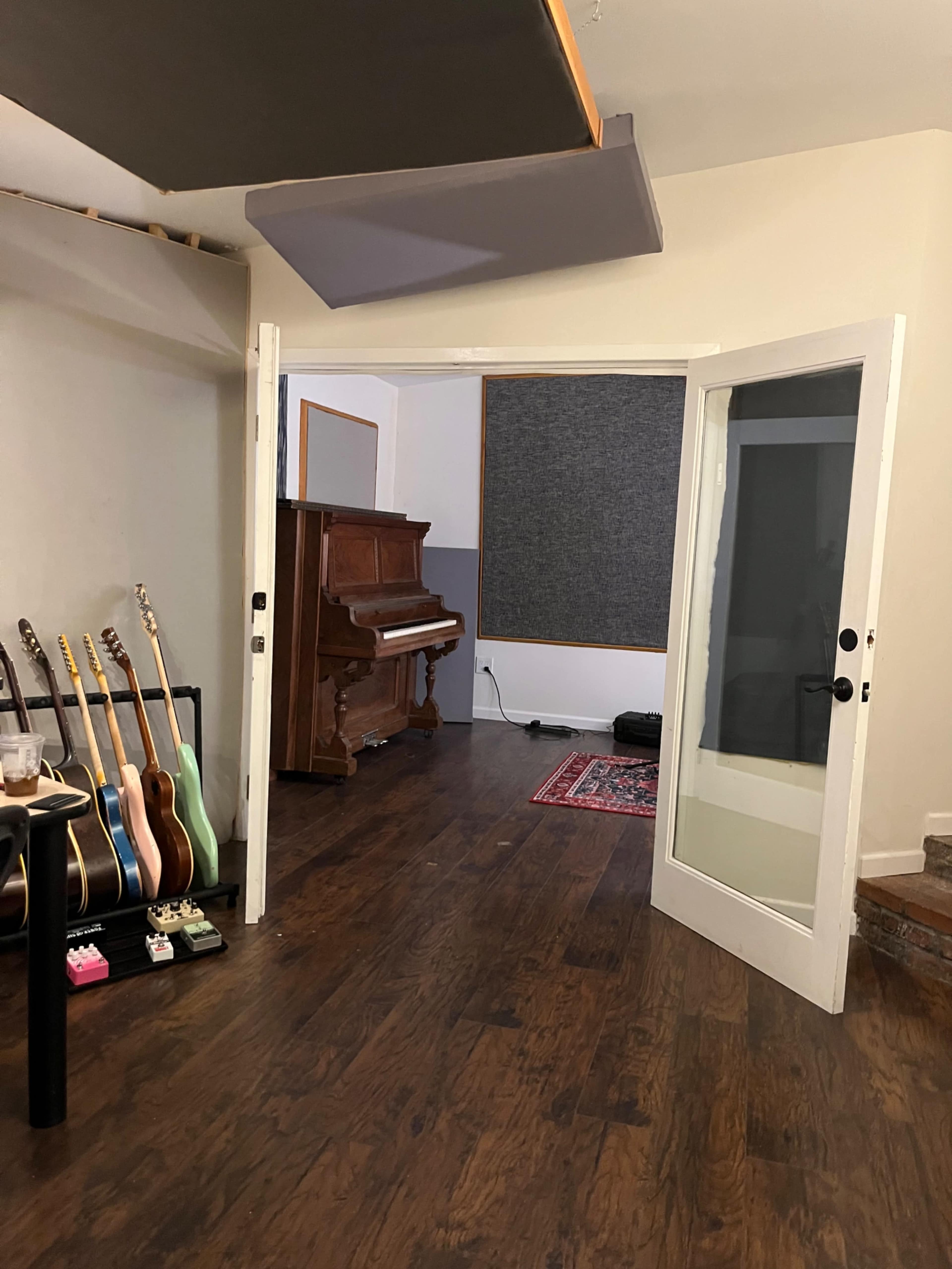 A room with an open doorway leading to a space that contains a vintage piano, several guitars on stands, and sound-absorbing panels on the walls.