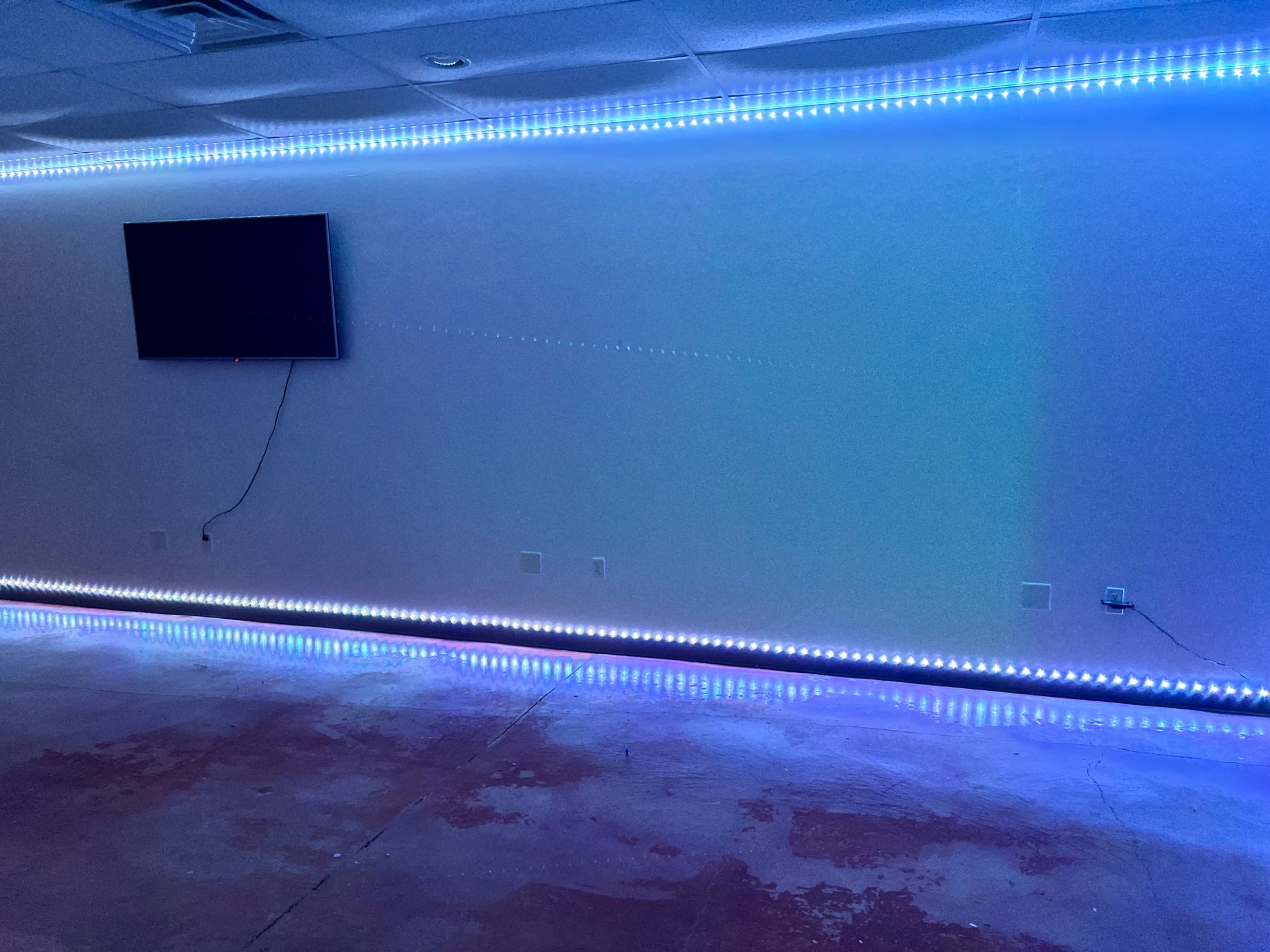 The image shows a room with a bare concrete floor and LED strip lighting along the perimeter, with a wall-mounted television on one side.