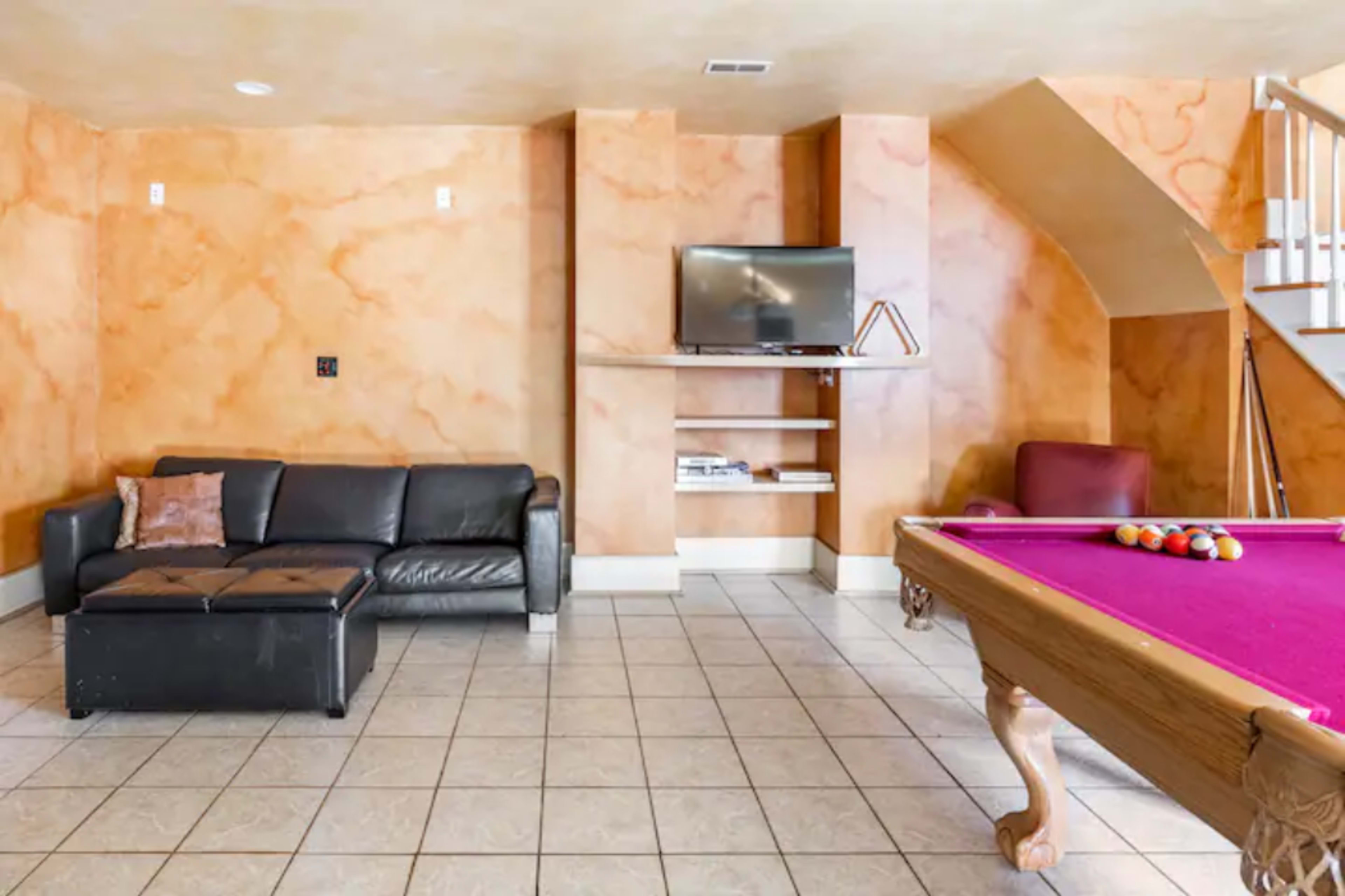 The room features a leather couch, a pool table with a purple felt surface, and a wall-mounted TV, all set against orange-painted walls.