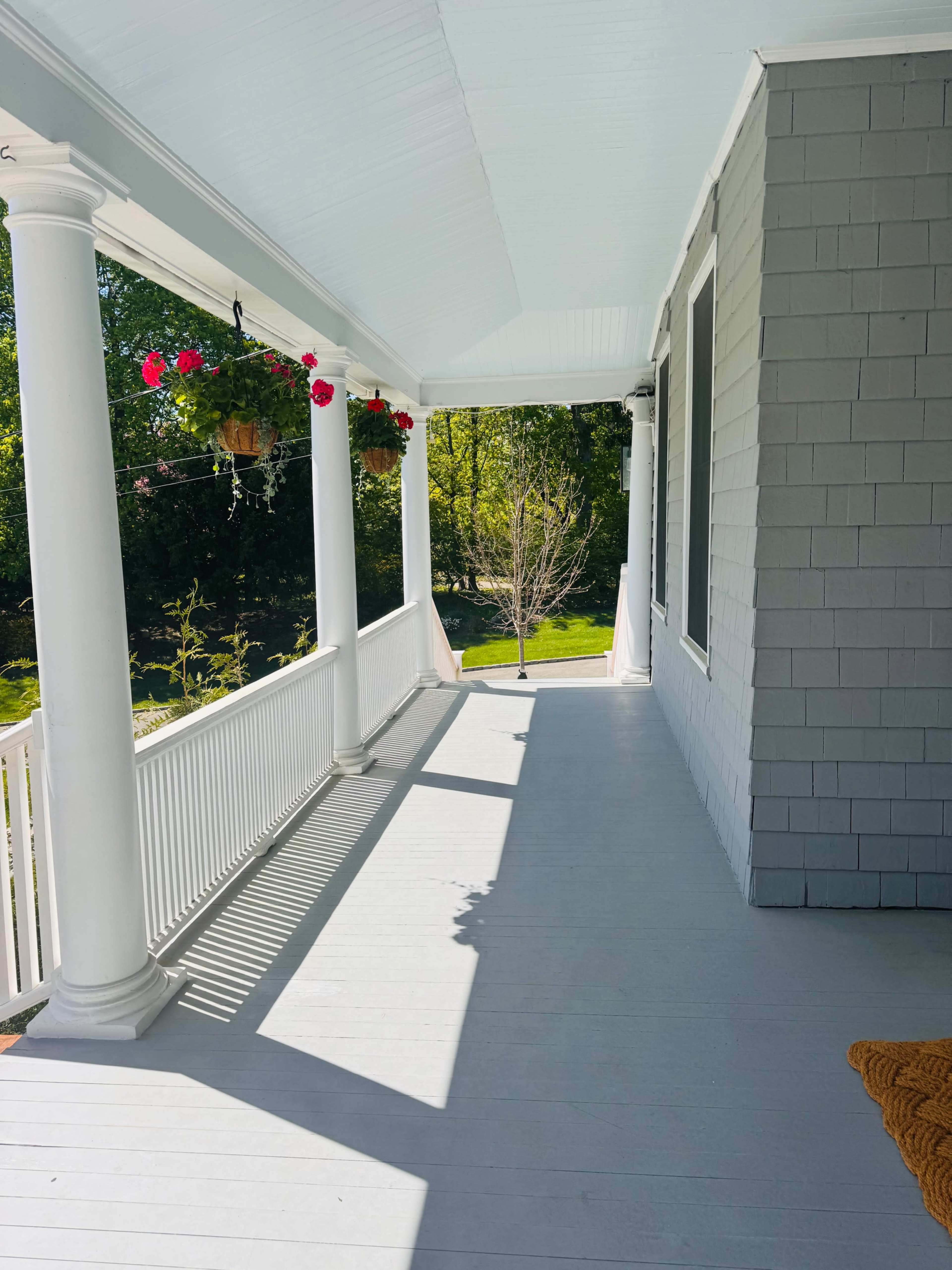 The image shows a bright, covered porch with white columns, featuring hanging flower pots and a view of a green yard.