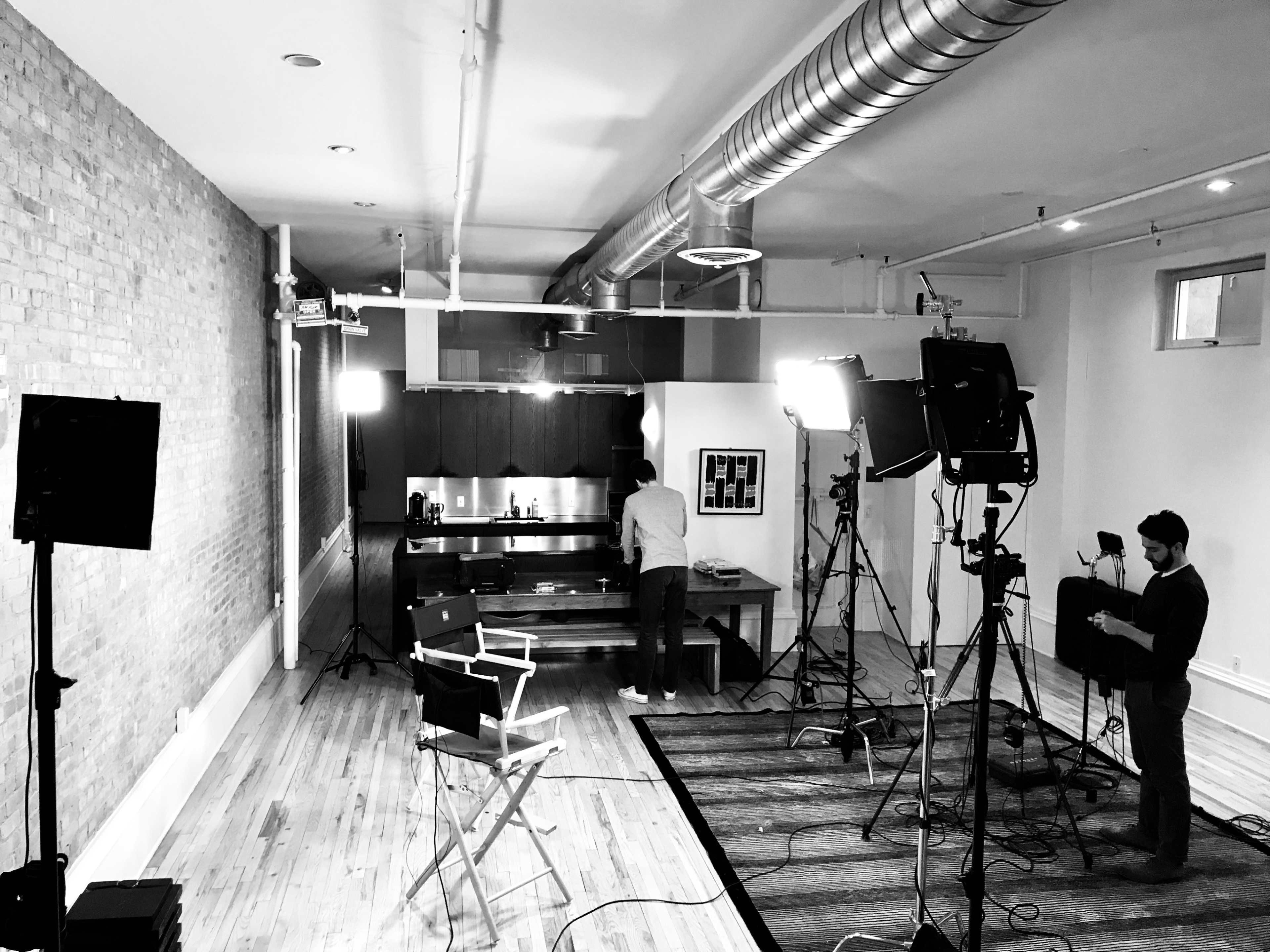 A spacious kitchen area is set up for a video shoot, with multiple cameras and lights positioned around a central wooden table where a person is preparing for filming.