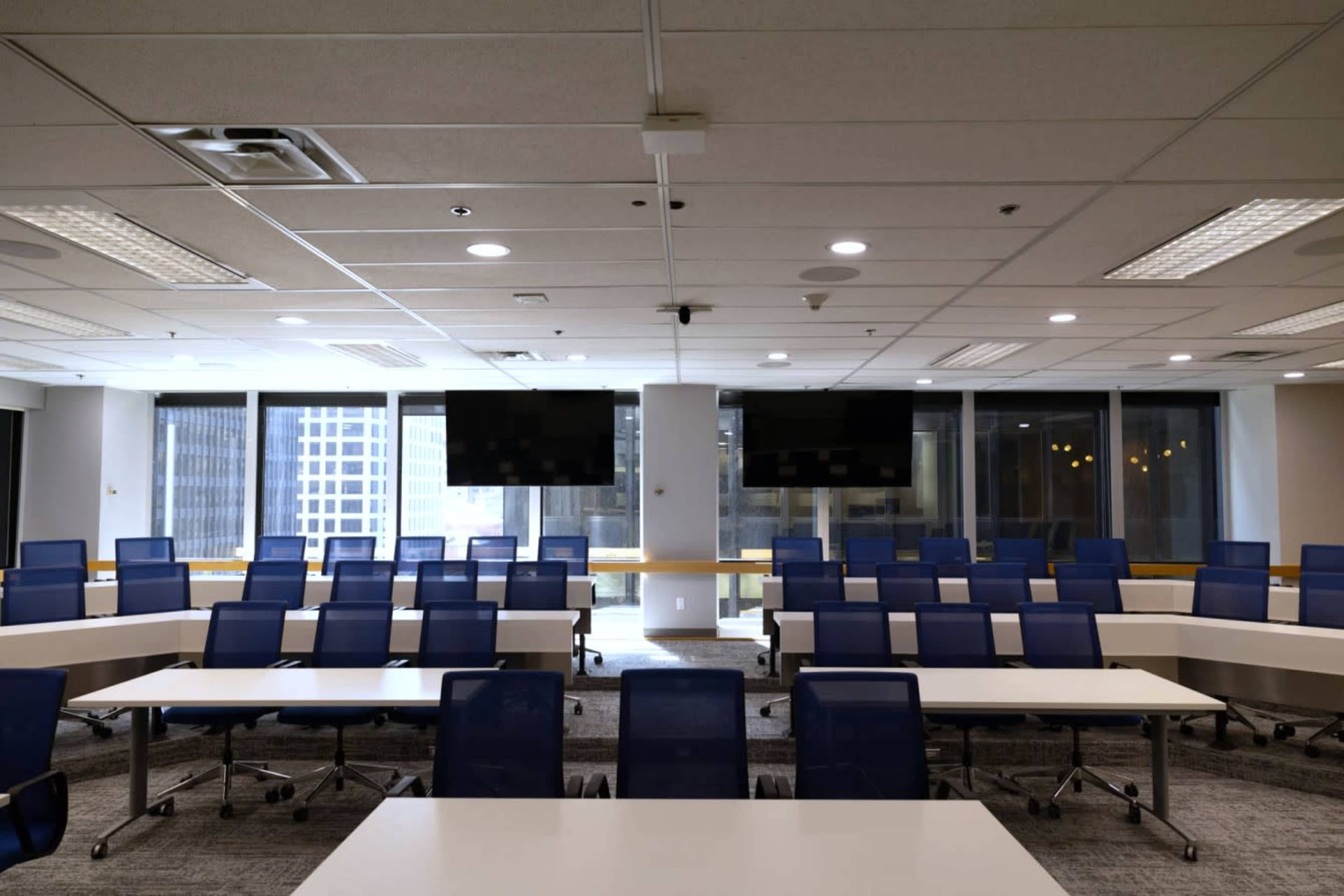 A modern conference room with rows of blue chairs and tables, large windows, and two flat-screen monitors on the wall.
