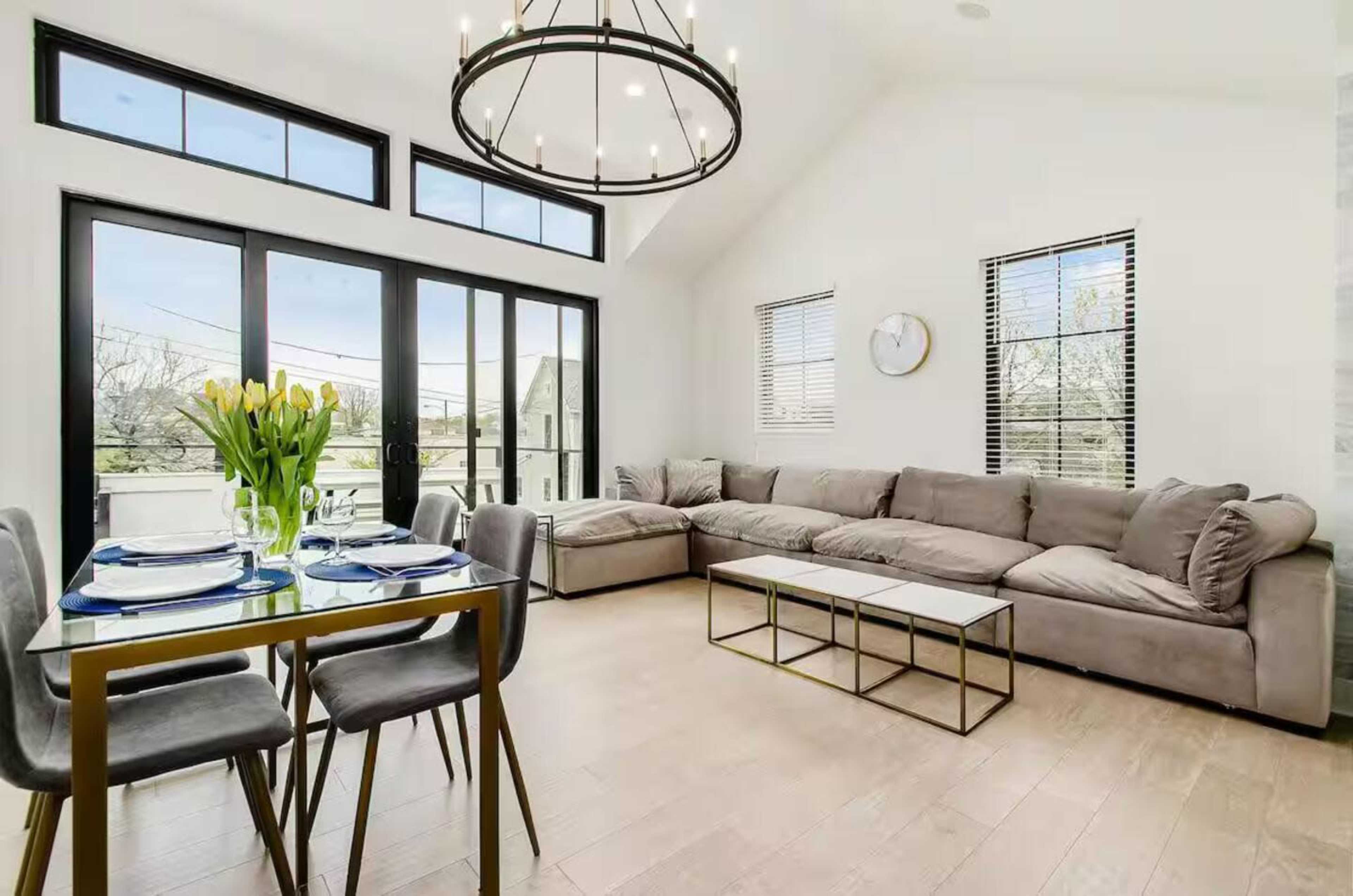 The image shows a modern living space featuring a sectional sofa, a dining table with place settings, and large windows allowing natural light to enter.