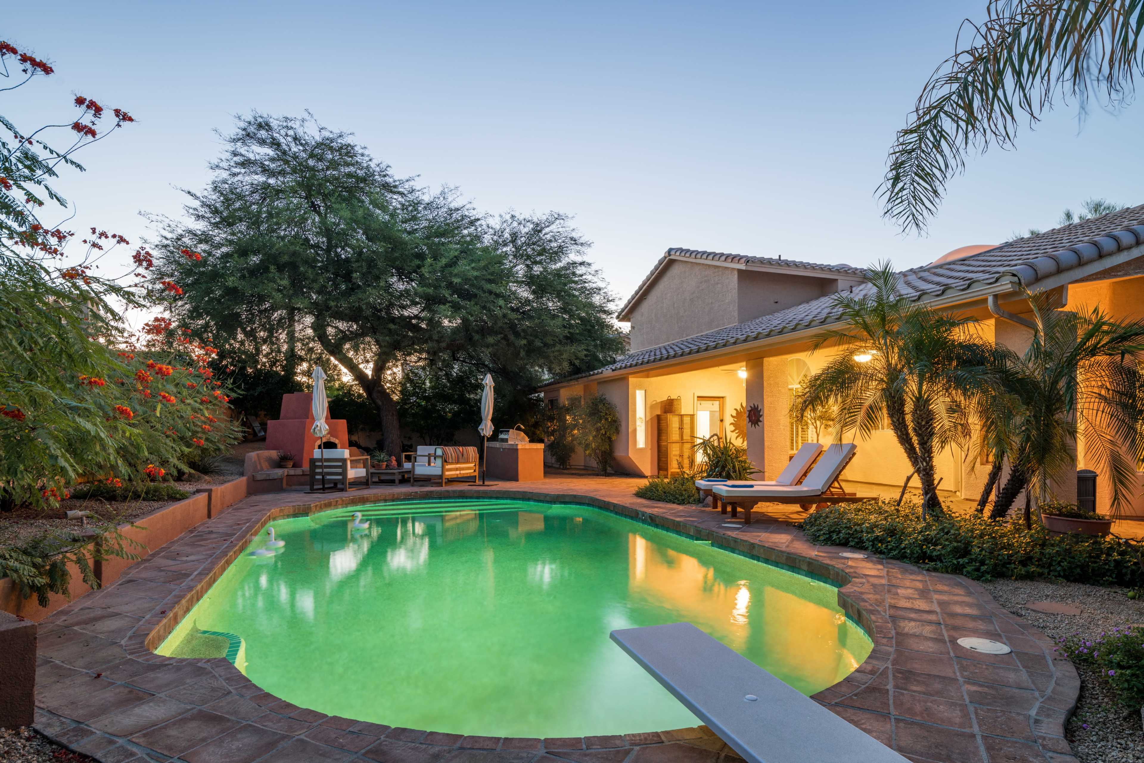 The image shows a tranquil backyard with a green swimming pool, lounging chairs, and surrounding tropical plants, all against a sunset sky.