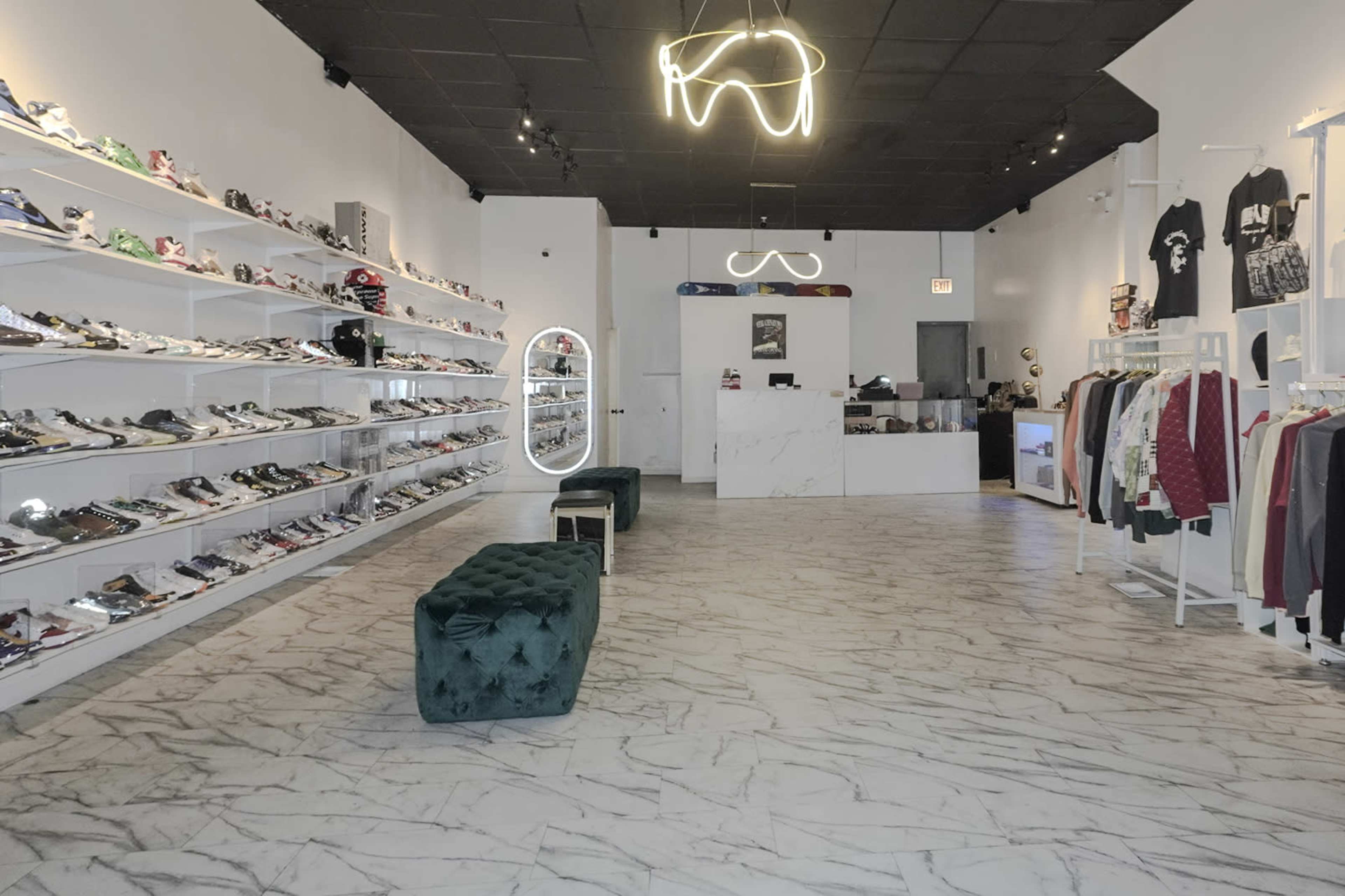 The interior of a retail store features shelves lined with various sneakers and clothing displays, with a minimalistic counter at the back and green velvet benches in the center.