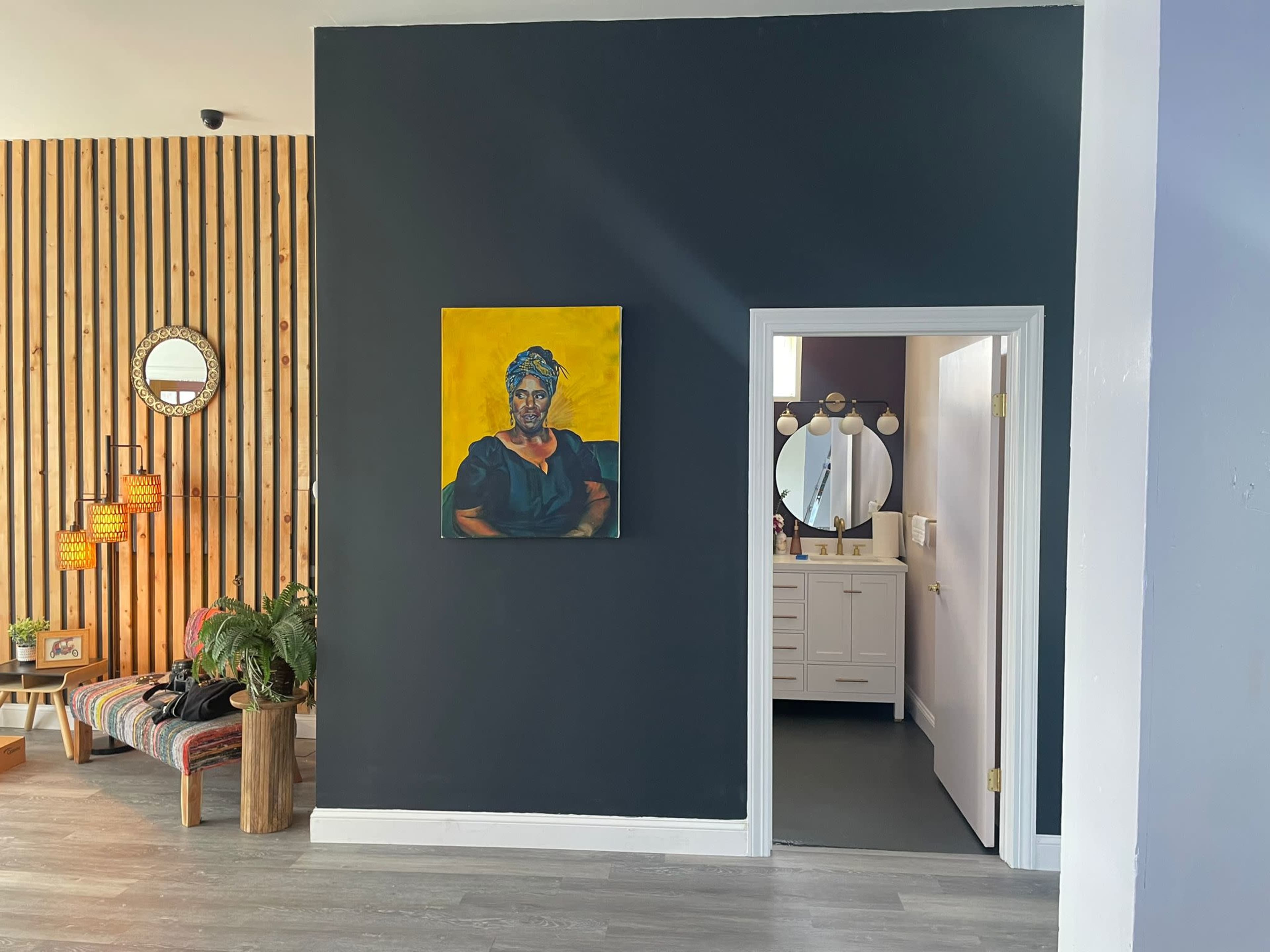 A modern interior featuring a dark wall with a portrait, a stylish seating area with a plant, and an adjoining room visible through an open doorway leading to a bathroom.