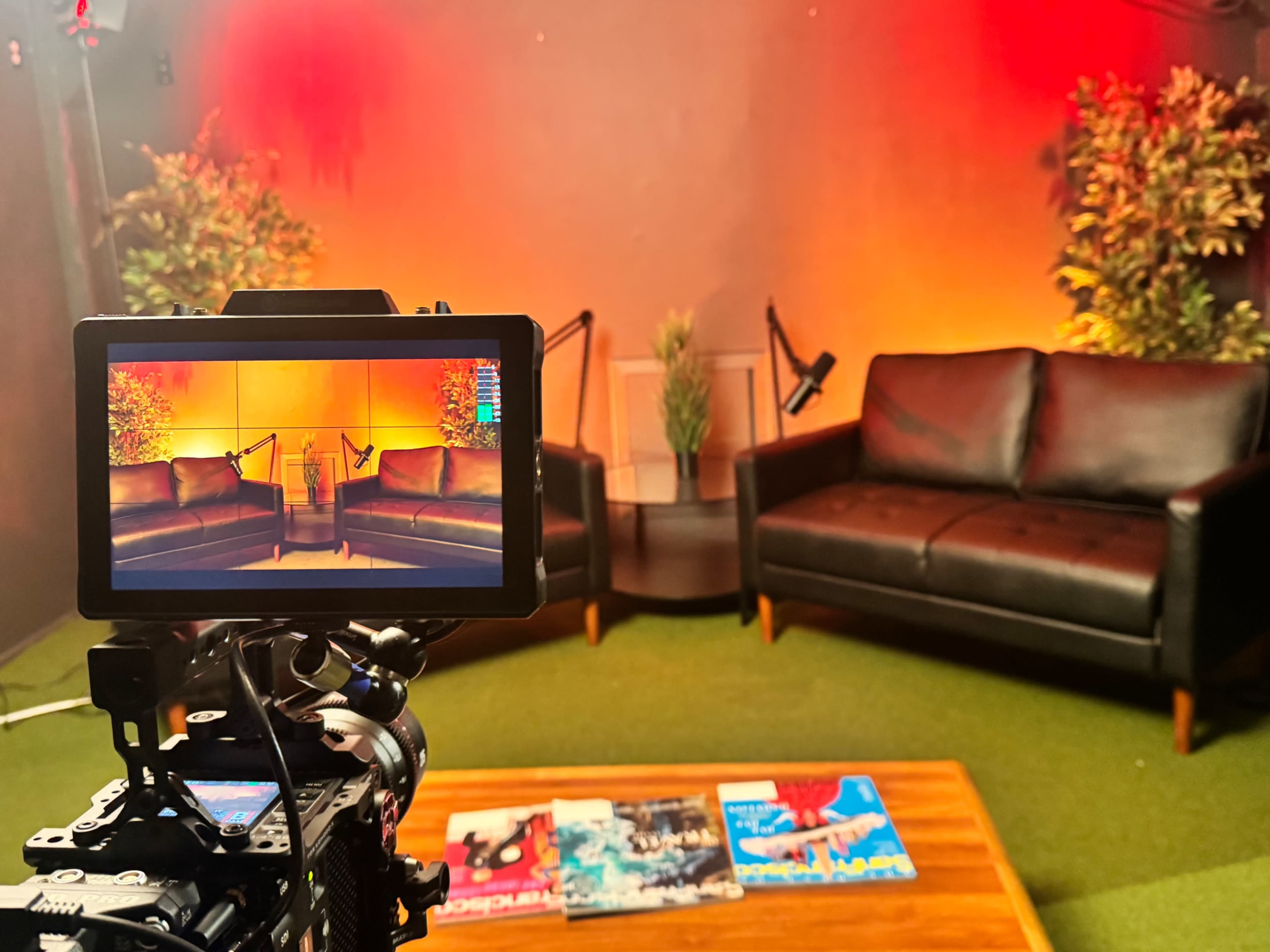 A camera on a tripod captures a view of a studio set featuring two black couches, decorative plants, and a wooden coffee table with magazines on it, all against a backdrop of orange lighting.