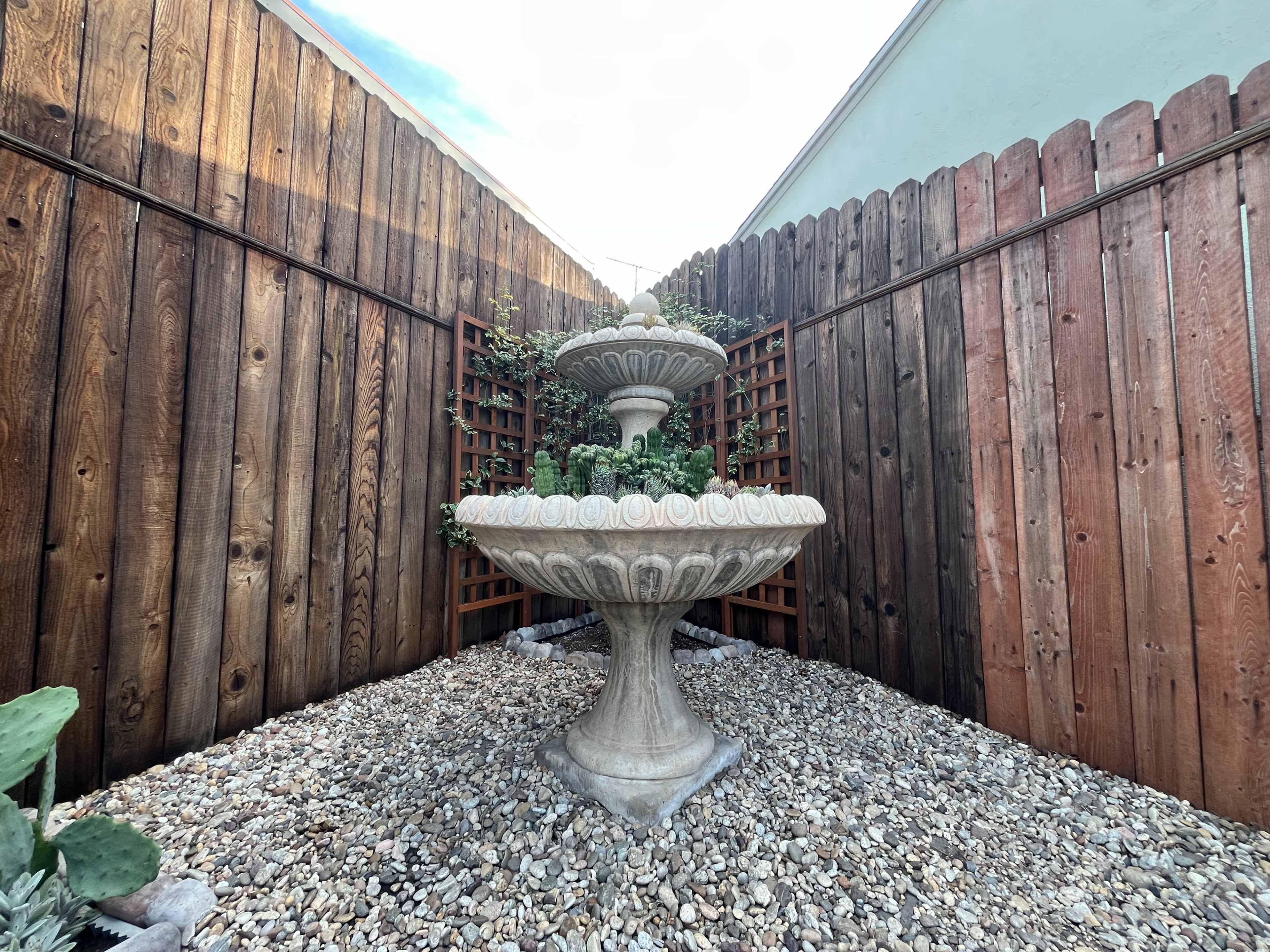 A two-tiered stone fountain is located in a gravel garden, surrounded by wooden fences and trellises with climbing plants.
