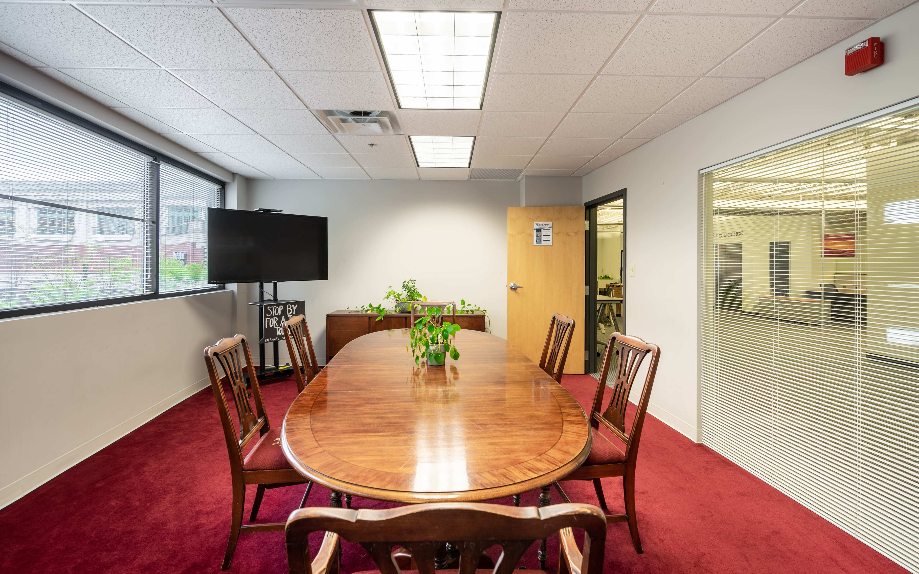 A large wooden table surrounded by chairs is set in a conference room with windows allowing natural light, featuring a television screen and glass-paneled doors.