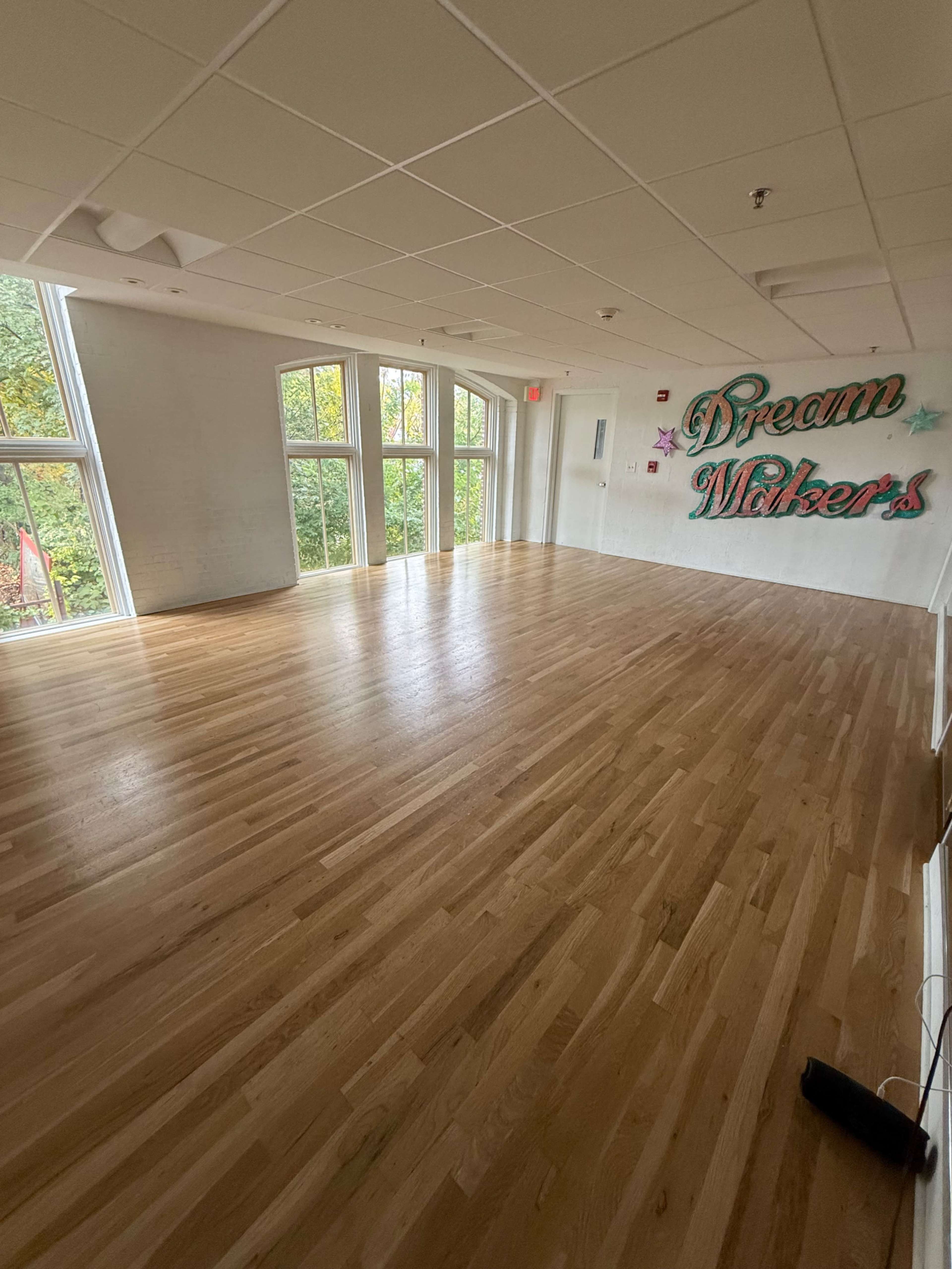 The image shows an empty wooden-floored room with large windows and a wall featuring the text "Dream Makers."