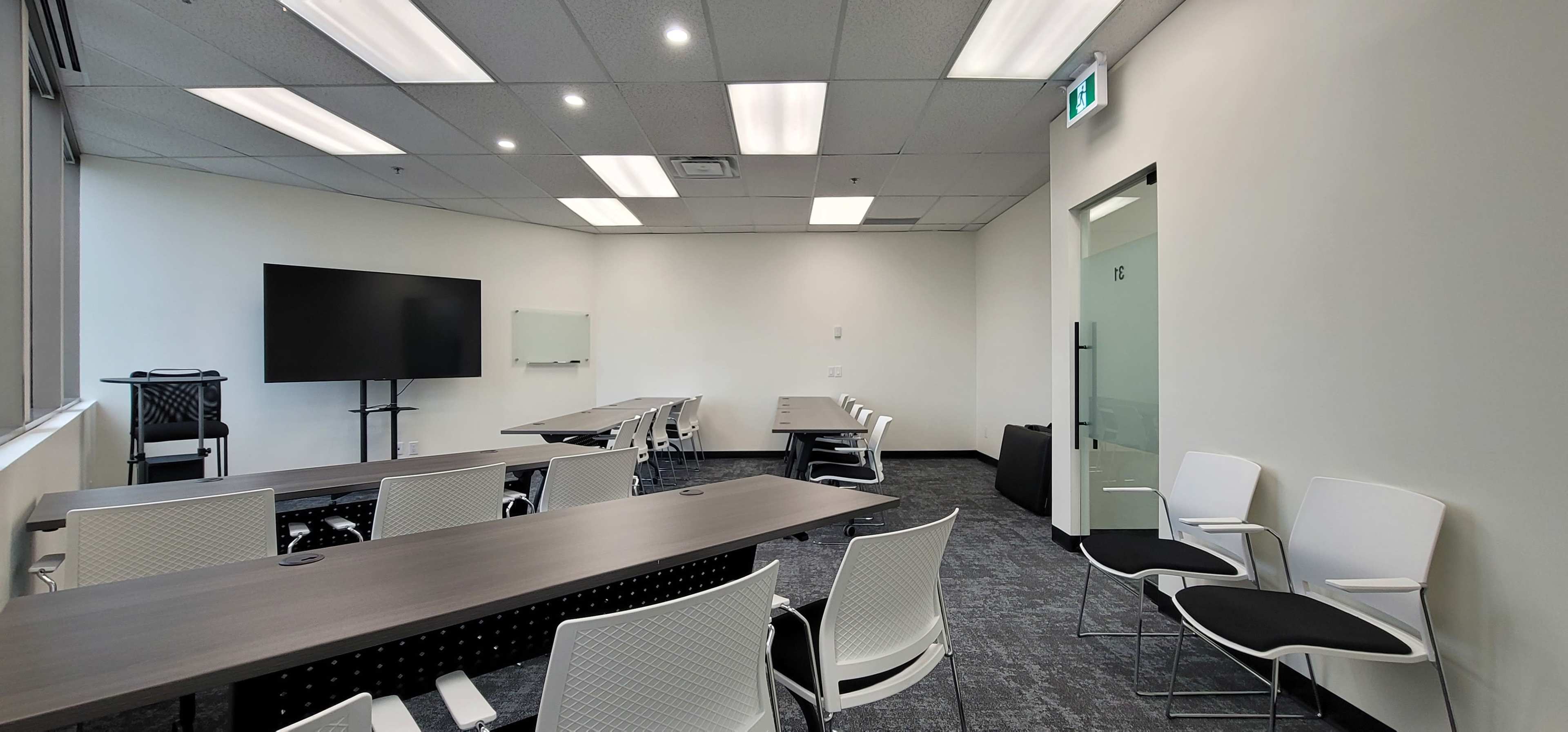 A meeting room with a large screen, whiteboard, and several white chairs arranged around a long table.